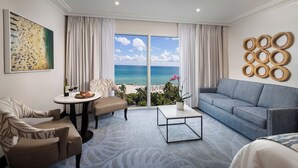Une chambre d’hôtel moderne dotée d’une grande fenêtre donnant sur la plage, d’un canapé, d’une table à manger avec une assiette de nourriture et d’un élément décoratif mural.