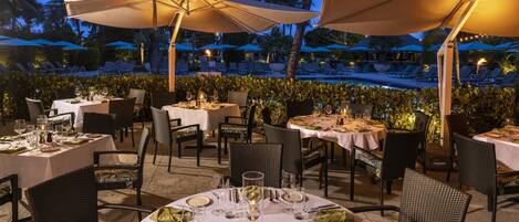 An outdoor dining area with round tables, chairs, and umbrellas, overlooking a pool and palm trees.