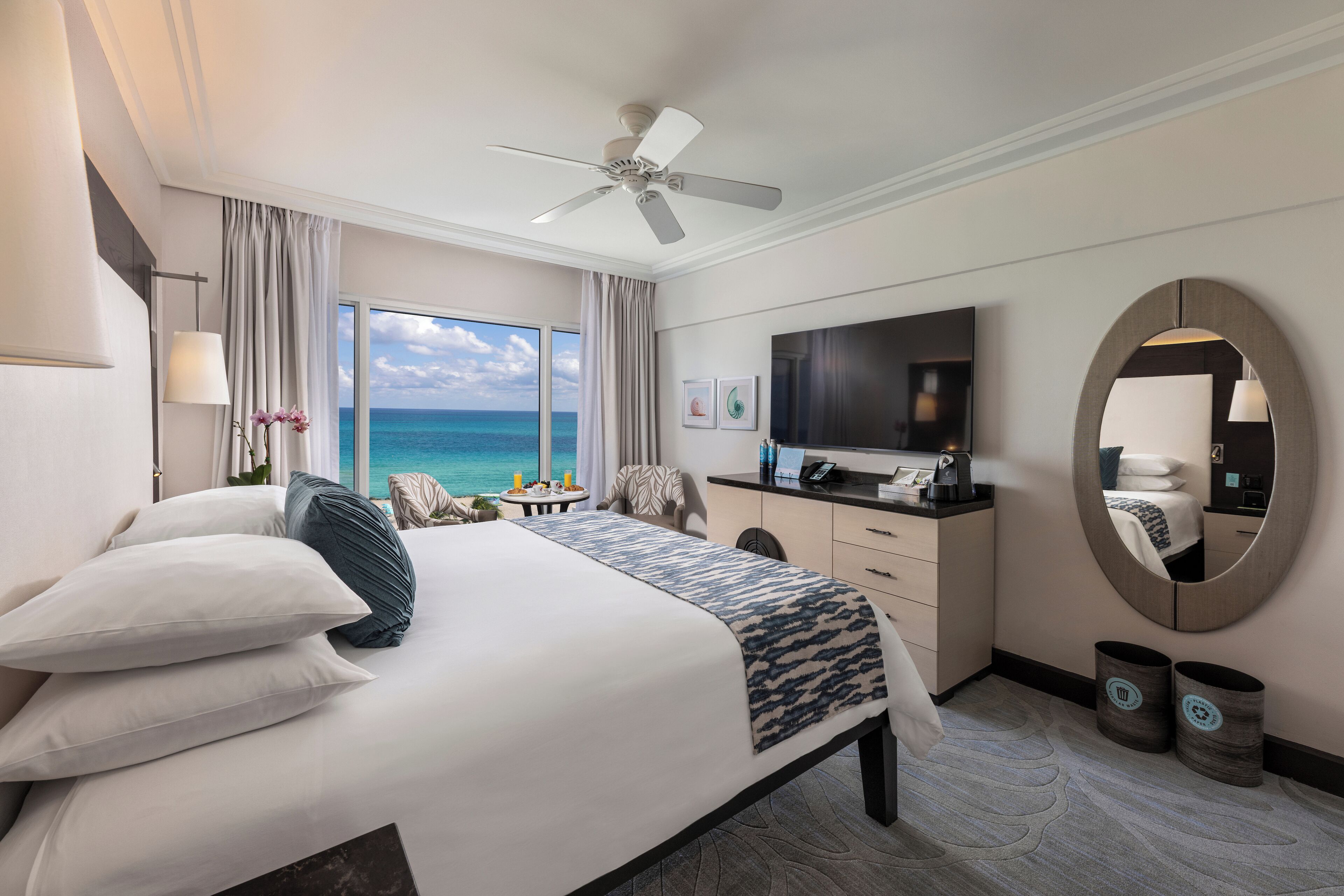 Une chambre d’hôtel comprenant un lit, un bureau, une télévision, une grande fenêtre donnant sur l’océan, un ventilateur de plafond et un miroir rond.