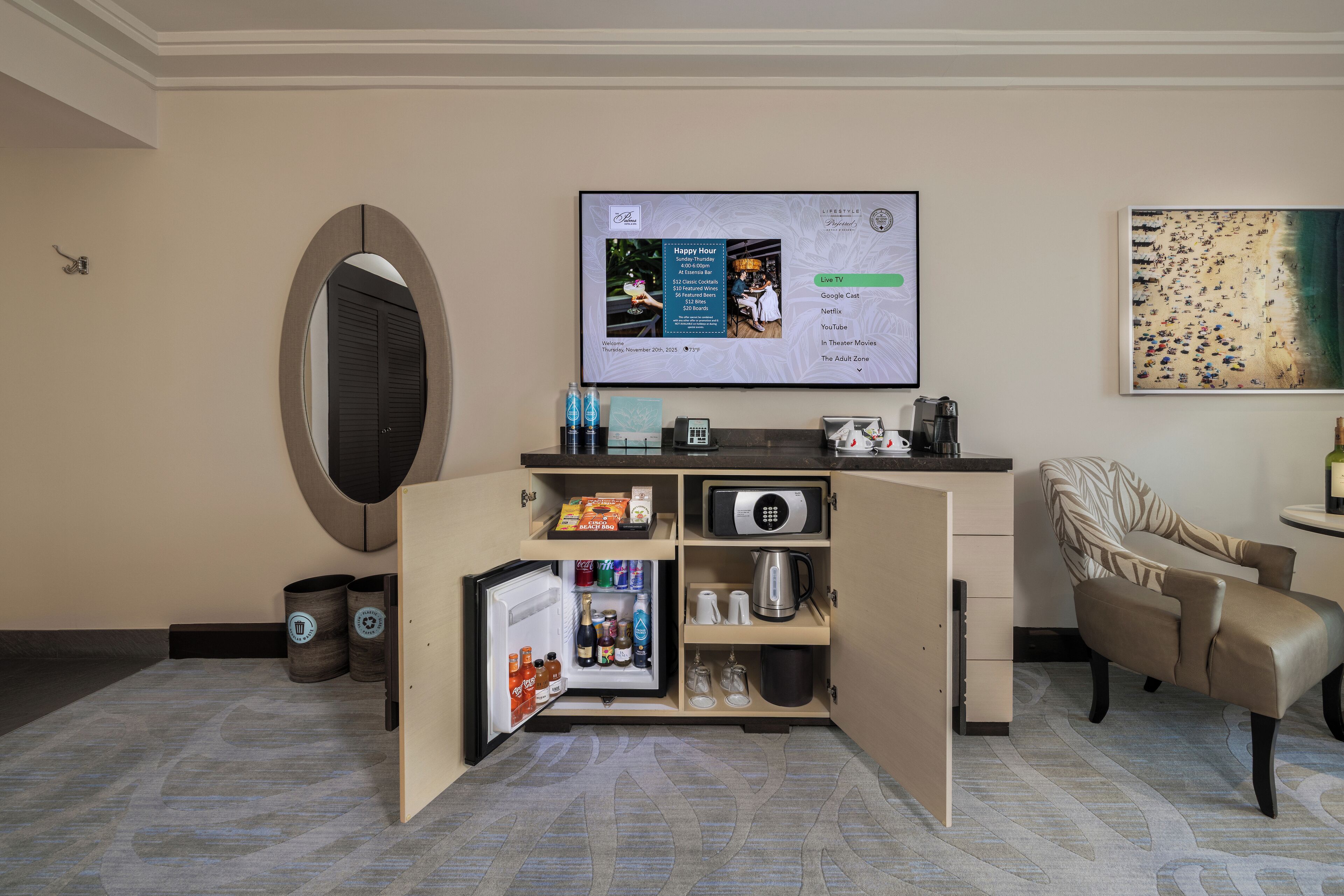 Quarto de hotel pequeno com minibar, televisão de tela plana exibindo interface de serviço de streaming, micro-ondas e área de estar com poltrona estampada.