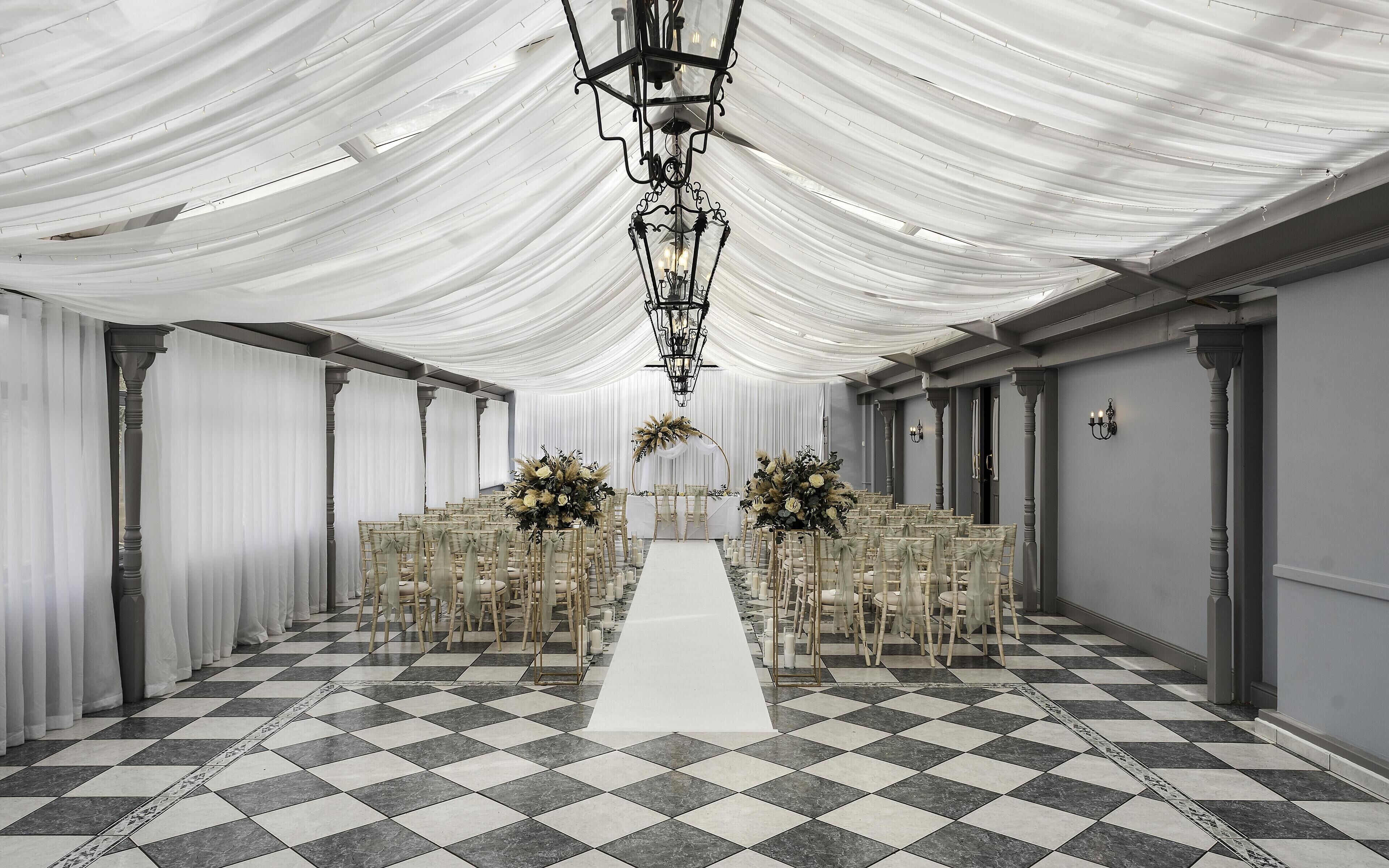 A venue with a checkered floor, white draped ceiling, and rows of chairs with floral centerpieces.