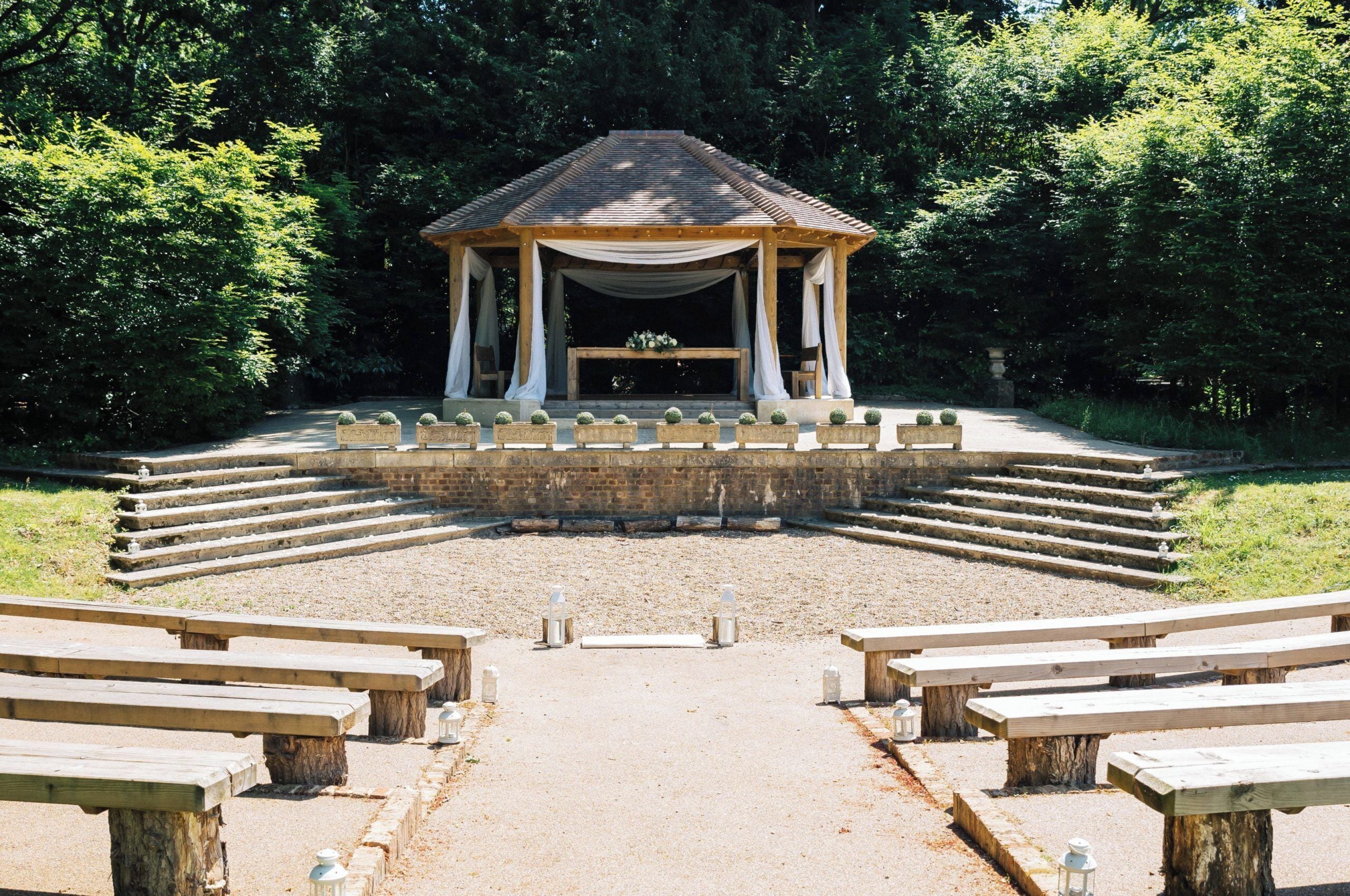 A gazebo with a tiled roof and white drapes, surrounded by a gravel area with benches.