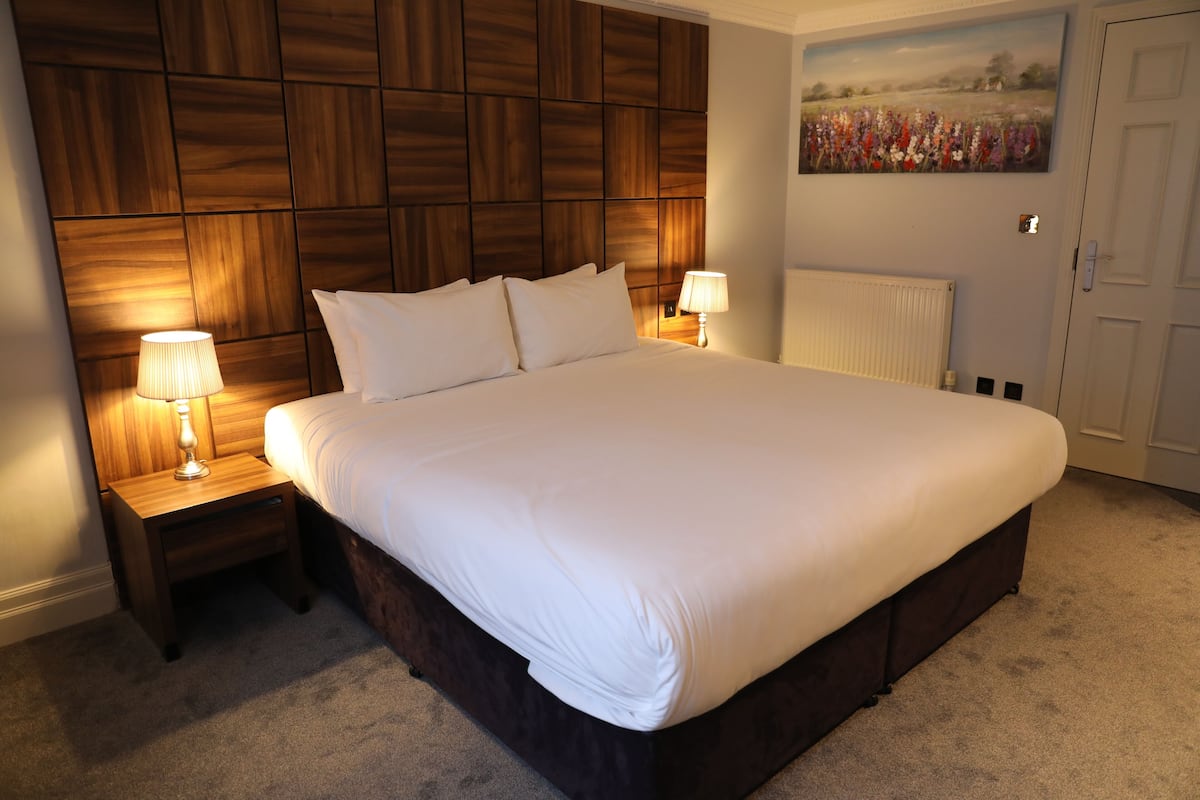 A bedroom with a large bed, wooden headboard, two bedside tables with lamps, a radiator, and a floral painting on the wall.