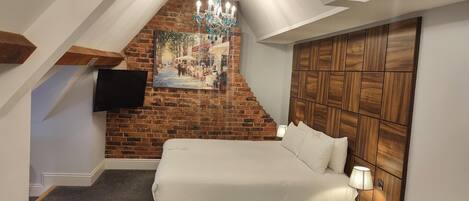 Un dormitorio con pared de ladrillo, un cuadro, cabecera de madera, cama y televisor montado en la pared.