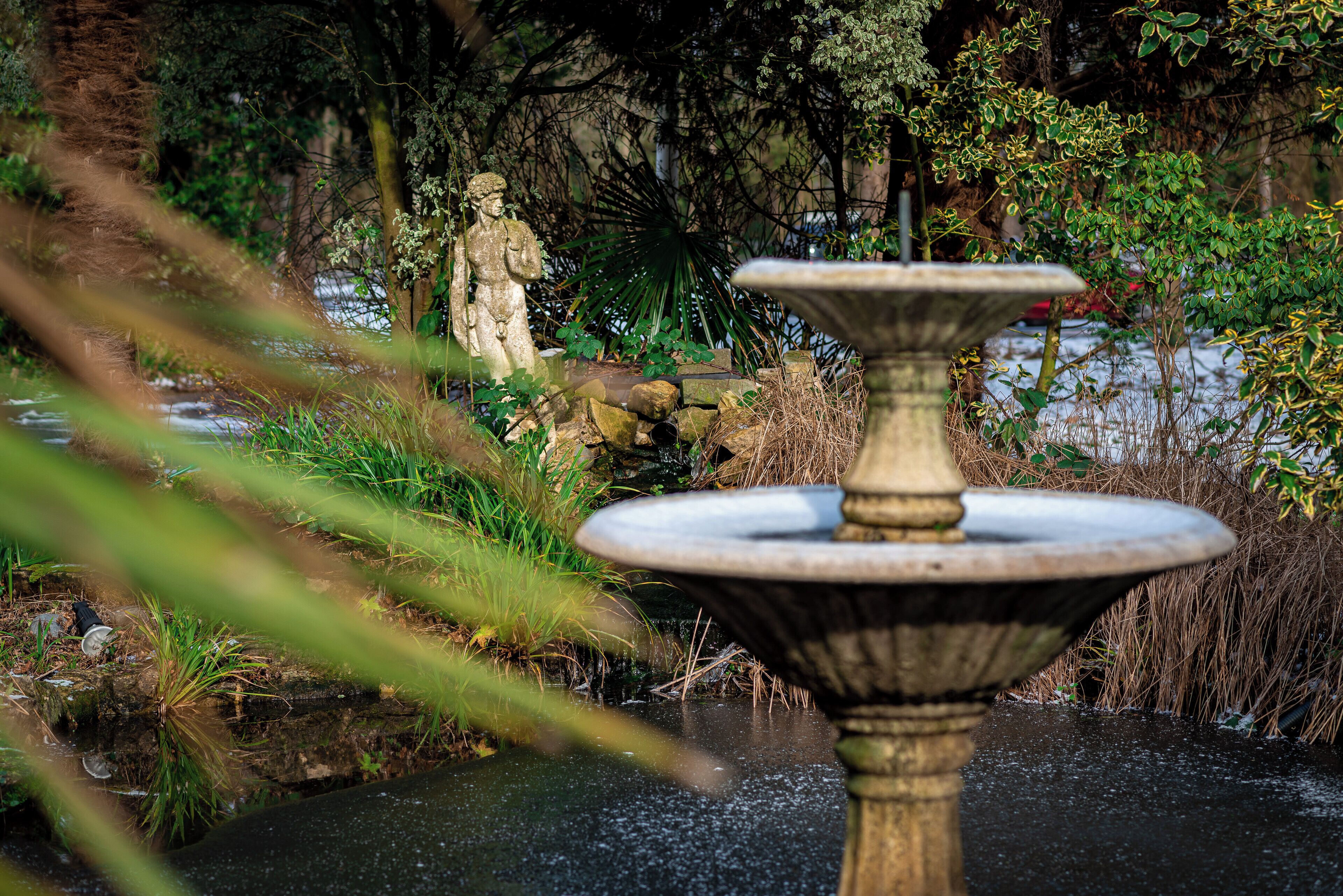 Une fontaine en pierre, avec une statue en arrière-plan, entourée de verdure et bordée d’un plan d’eau.