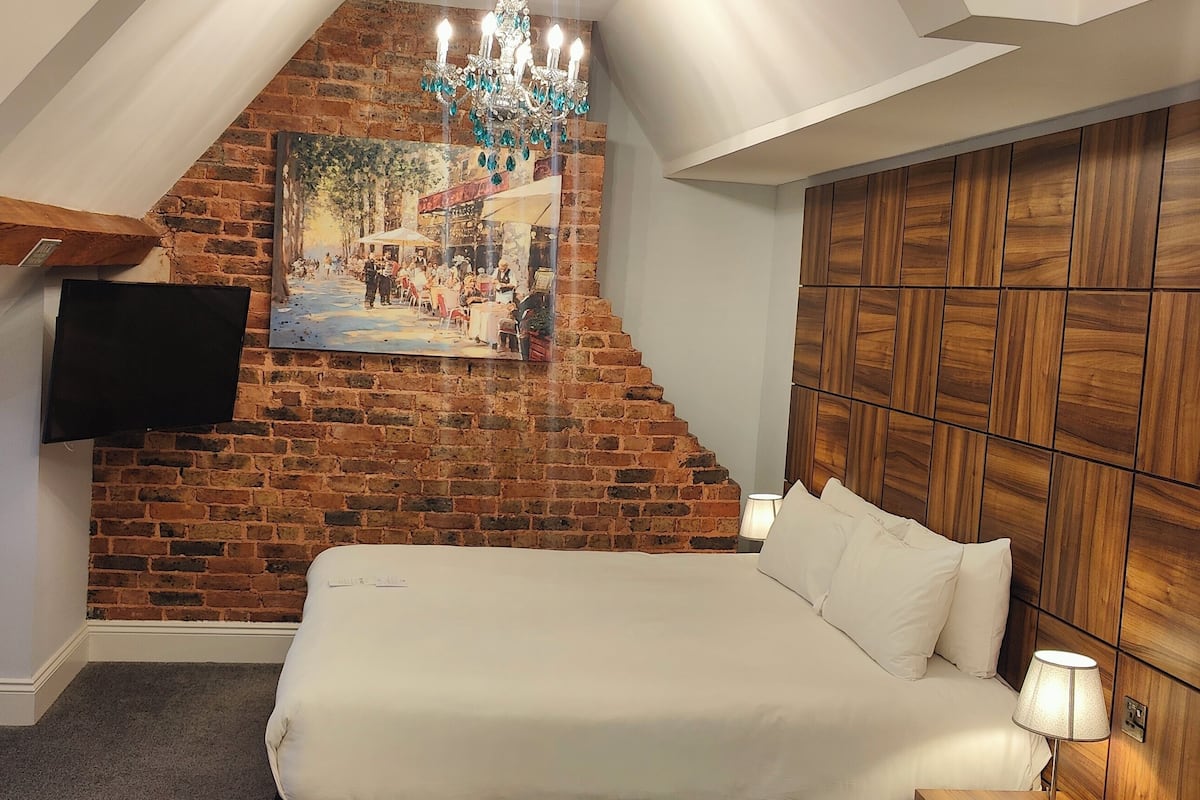 A bedroom with a brick wall, a mounted TV, a wooden headboard, and a painting of a street scene.