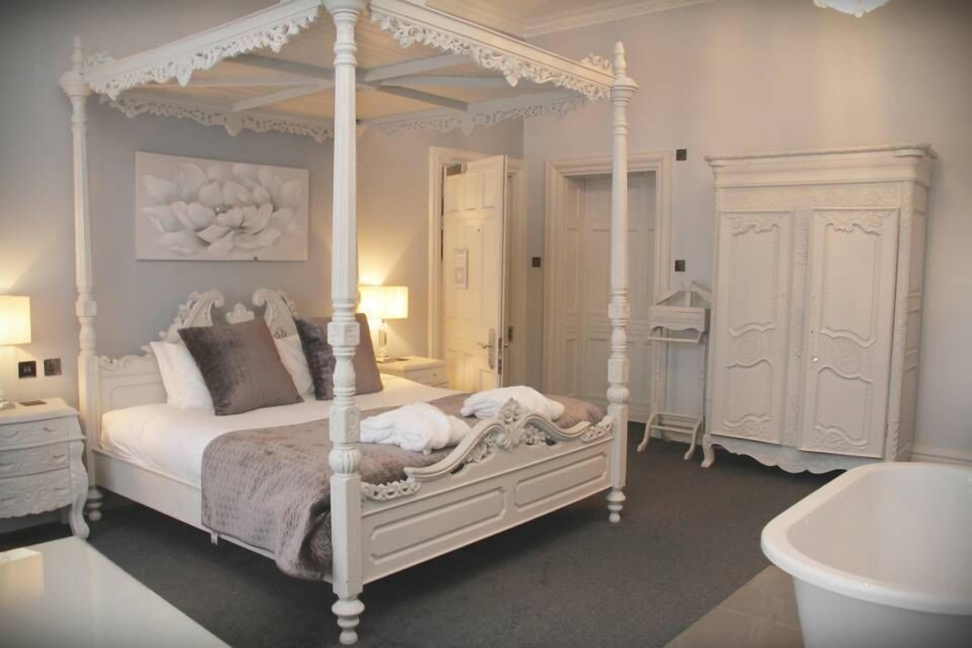 A four-poster canopy bed with a white finish, a bedside table with a lamp, and a wardrobe with a mirror.