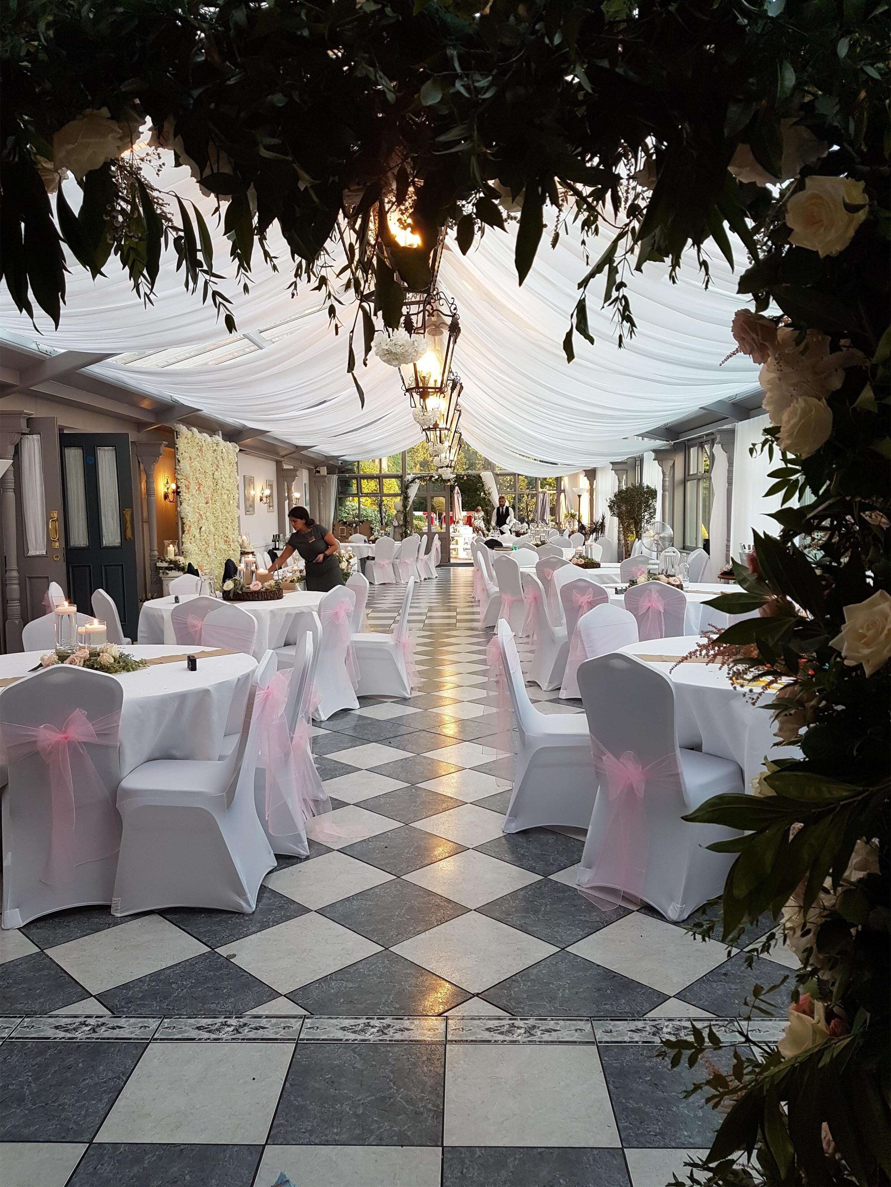 Un salón de banquetes con manteles blancos, sillas adornadas con cintas rosas y suelo a cuadros.