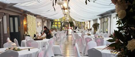 Un salón de banquetes con manteles blancos, sillas adornadas con cintas rosas y suelo a cuadros.