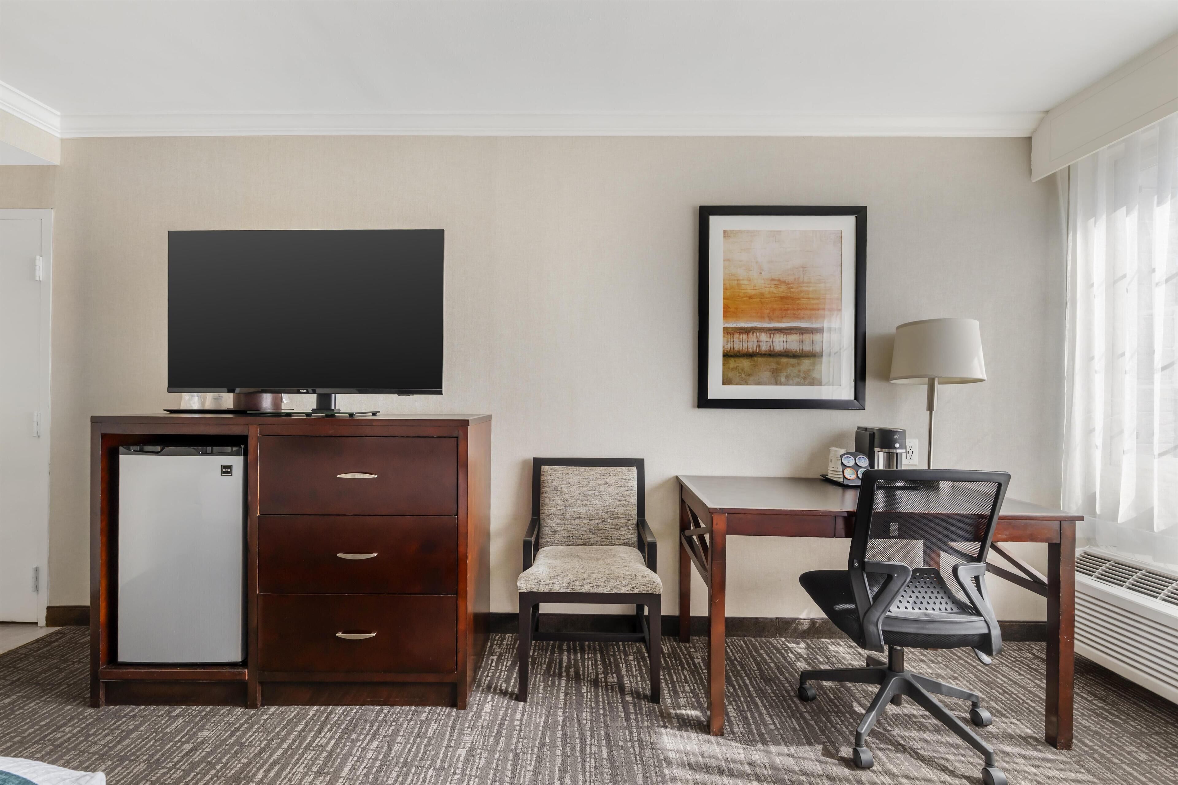 A hotel room with a flat-screen TV, a desk with a chair, a small refrigerator, a cabinet with drawers, a framed picture on the wall, and a window with curtains.
