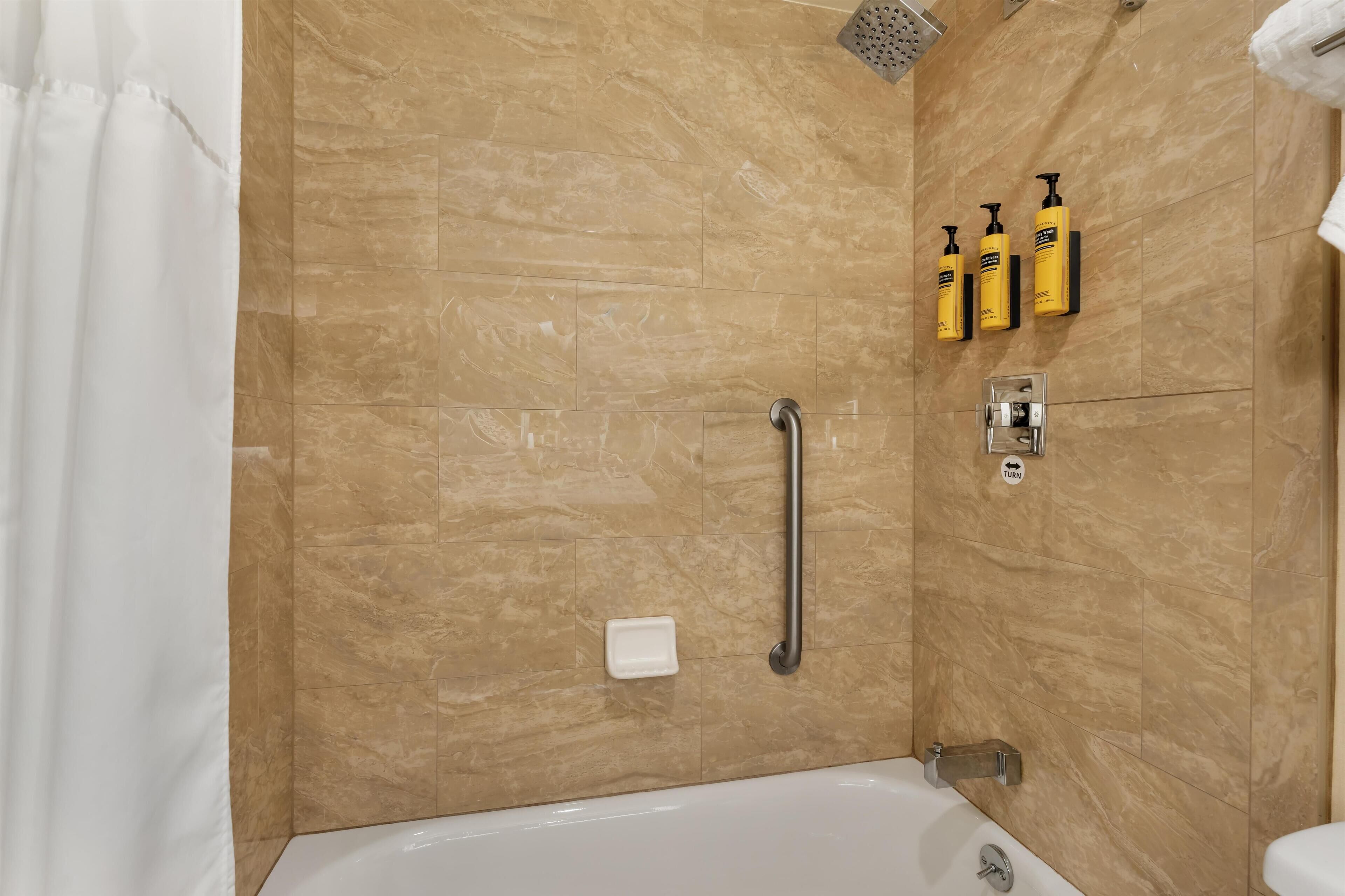 Un baño con bañera, ducha y barra de apoyo. Hay tres botellas de champú y acondicionador en la pared.