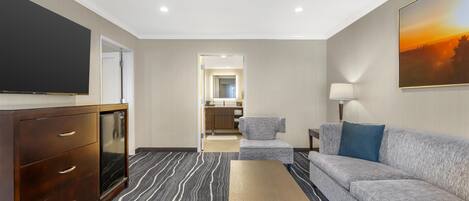 Habitación de hotel con televisor de pantalla plana, cómoda de madera, sofá gris, sillón pequeño, lámpara y un cuadro en la pared.