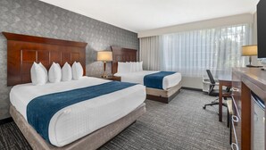A hotel room with two beds, a desk, a chair, and a large window with curtains.