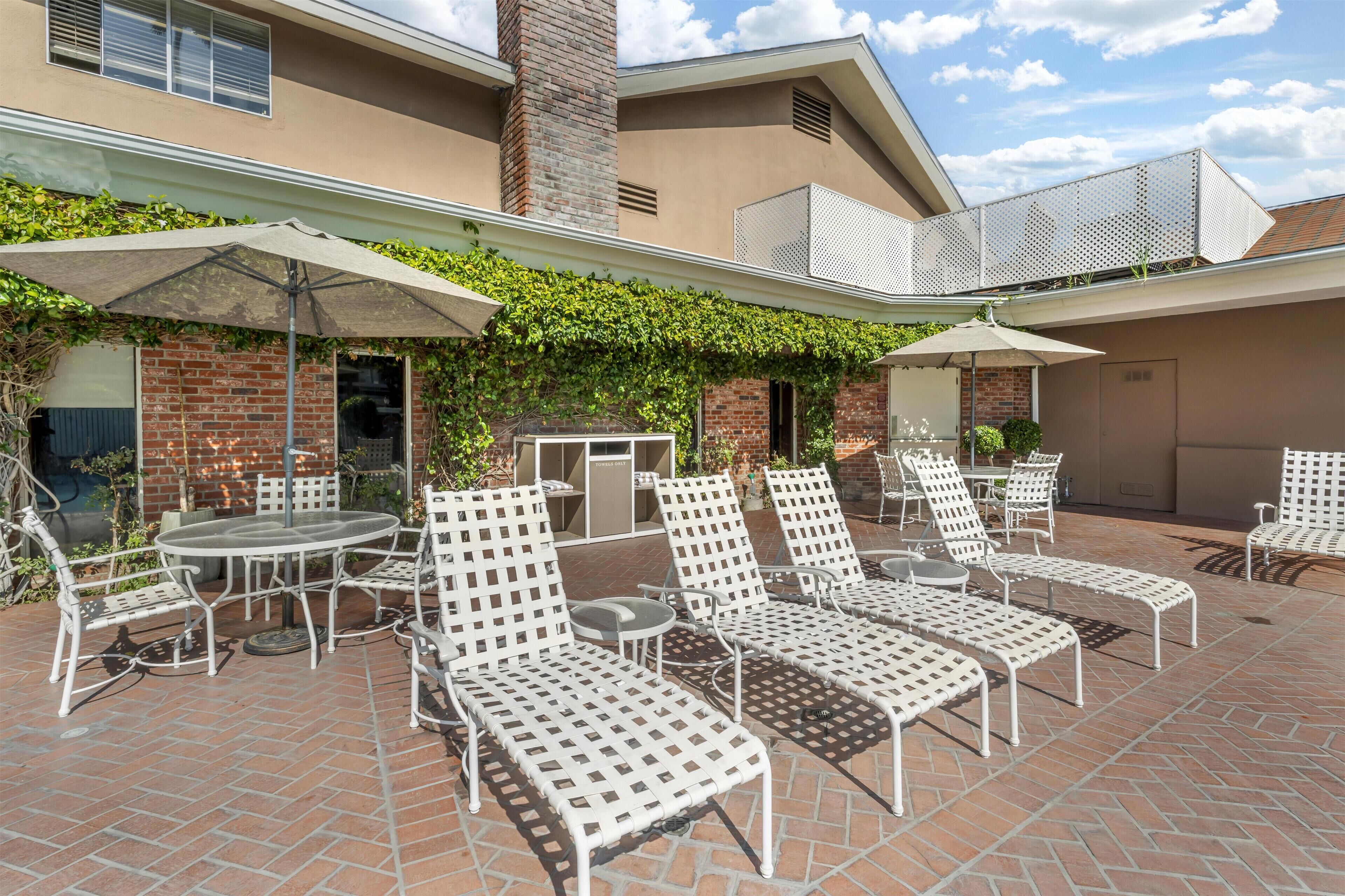 Une terrasse extérieure avec des chaises et des tables en métal blanc, un mur de briques et un espace abrité sous un parasol blanc.