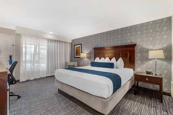 A hotel room with a large bed, a desk, a chair, a nightstand, and a wall-mounted lamp.