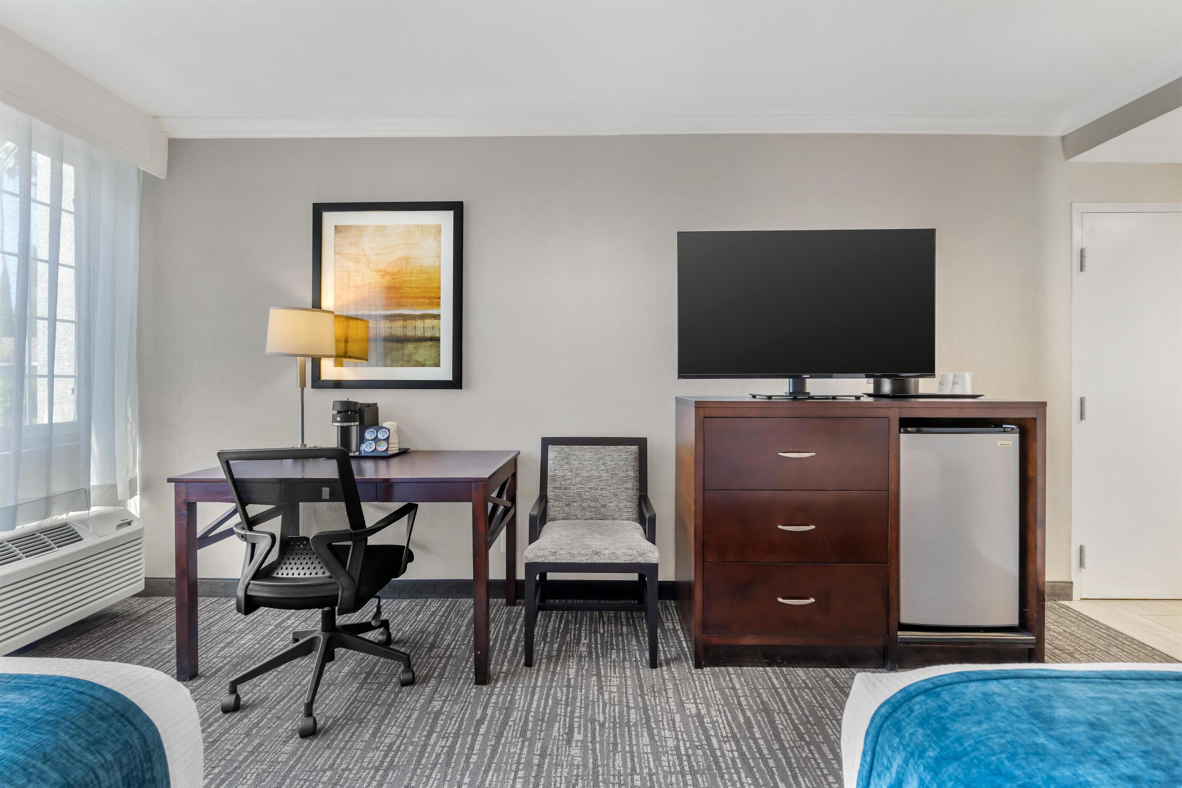 A hotel room with a bed, a desk with a chair, a television, a refrigerator, and a wall-mounted TV.