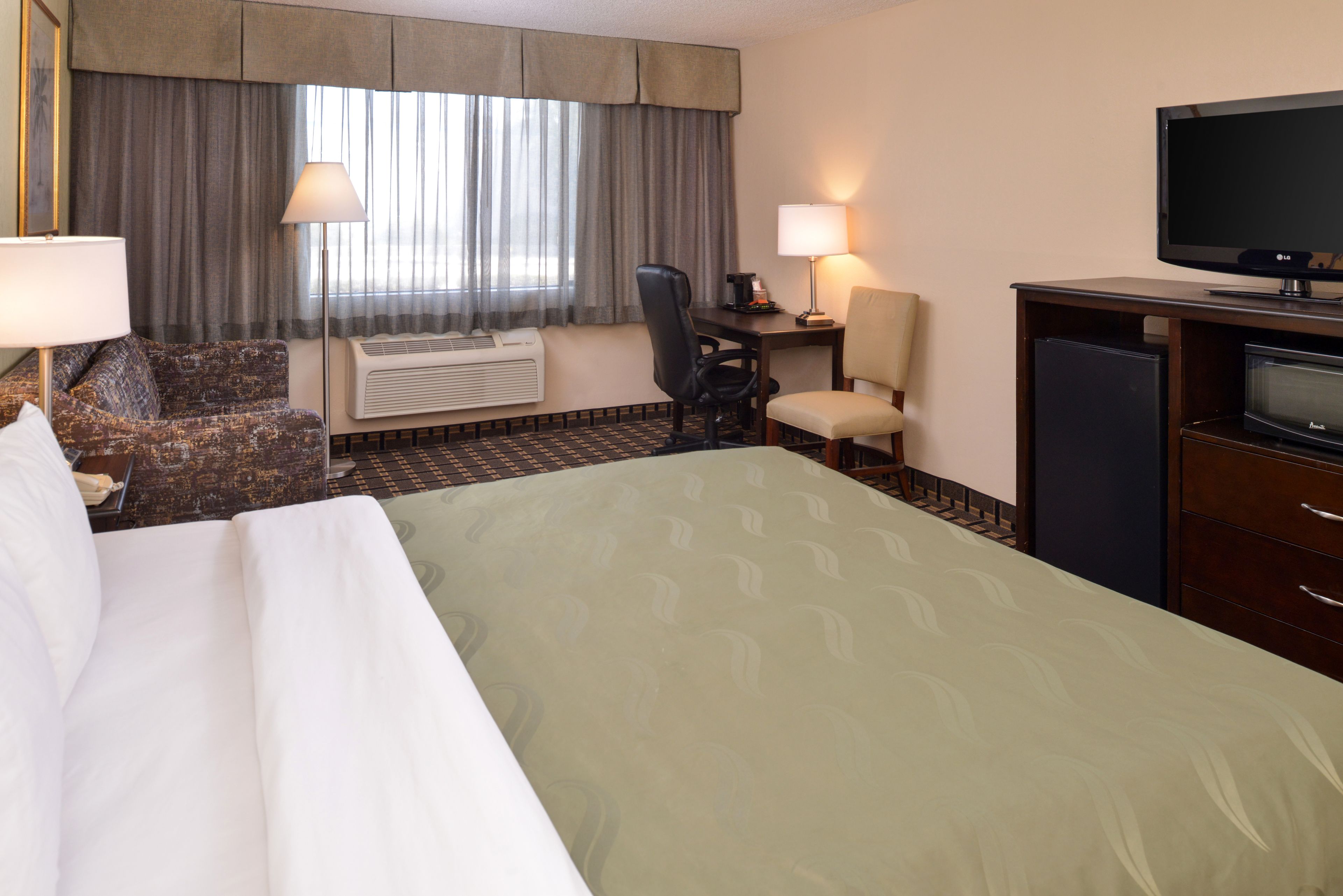 A hotel room with a large bed, two armchairs, a desk with a computer, a television, and a window with curtains.