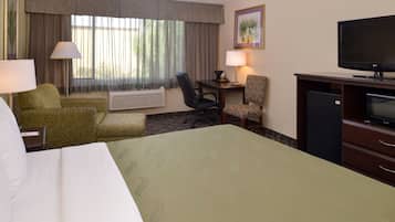 Quarto de hotel com uma cama grande, duas poltronas, uma mesa de trabalho com luminária, televisão e janela com cortinas.