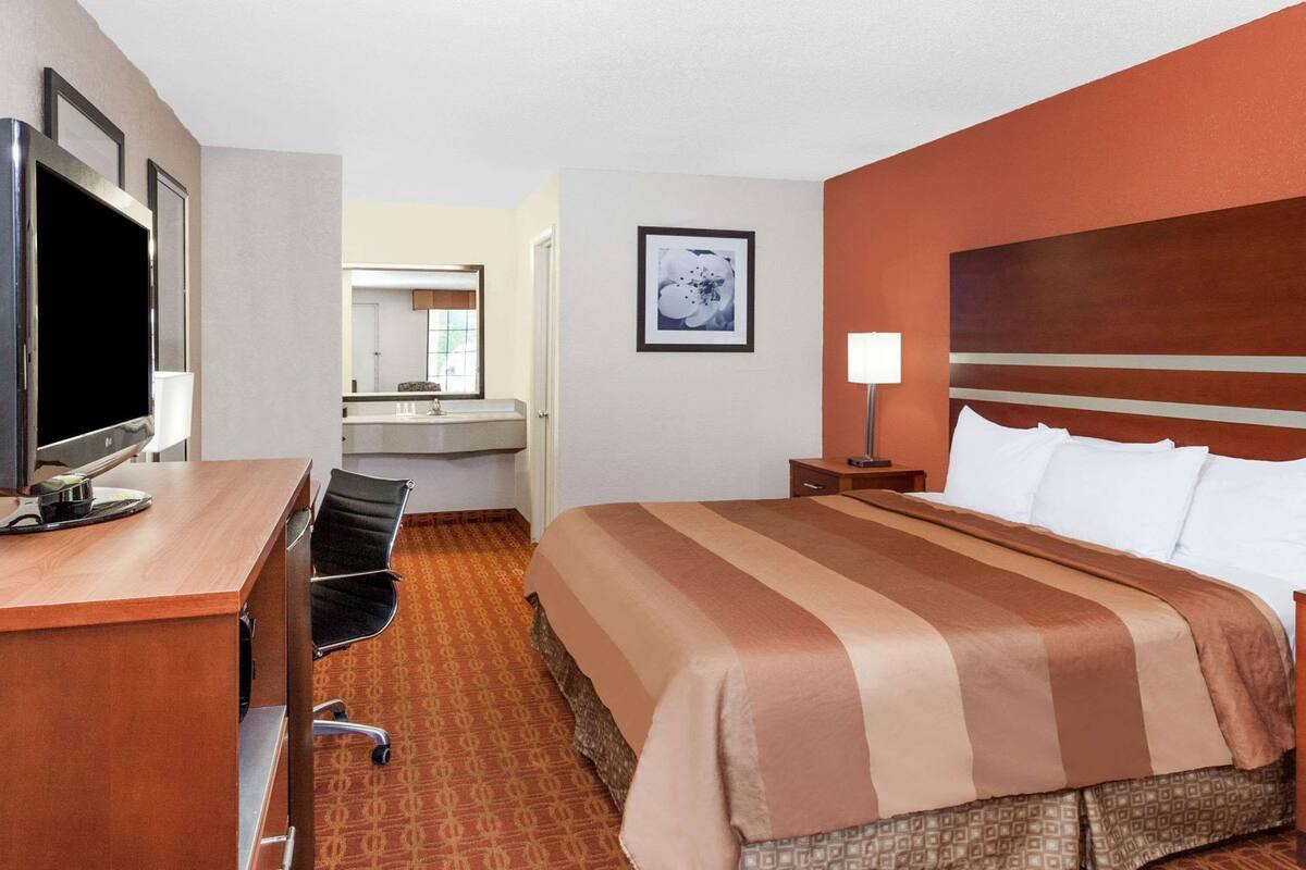 A hotel room with a bed, a desk with a TV, a chair, a bedside table with a lamp, and a bathroom visible through an open door.