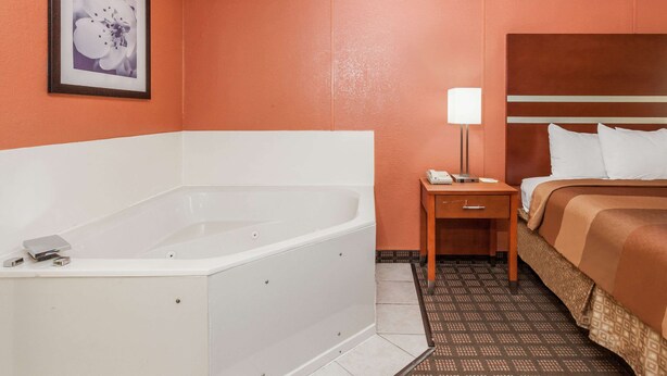 A hotel room with a large bathtub, a bedside table with a lamp, and a bed with pillows. - Days Inn by Wyndham Dalton (Dalton)