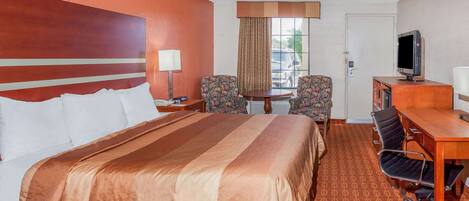 A hotel room with a large bed, two armchairs, a desk with a computer, and a television.