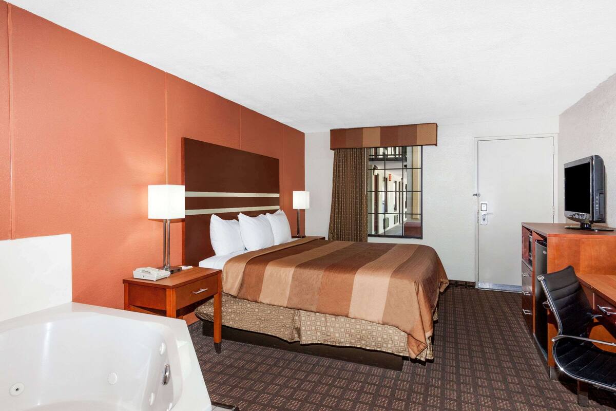 A hotel room with a large bed, a desk with a television, a desk lamp, a chair, a bathroom with a bathtub, and a window with curtains.