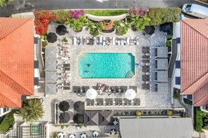 An aerial view of a resort with a central swimming pool, sun loungers, umbrellas, and a landscaped garden.