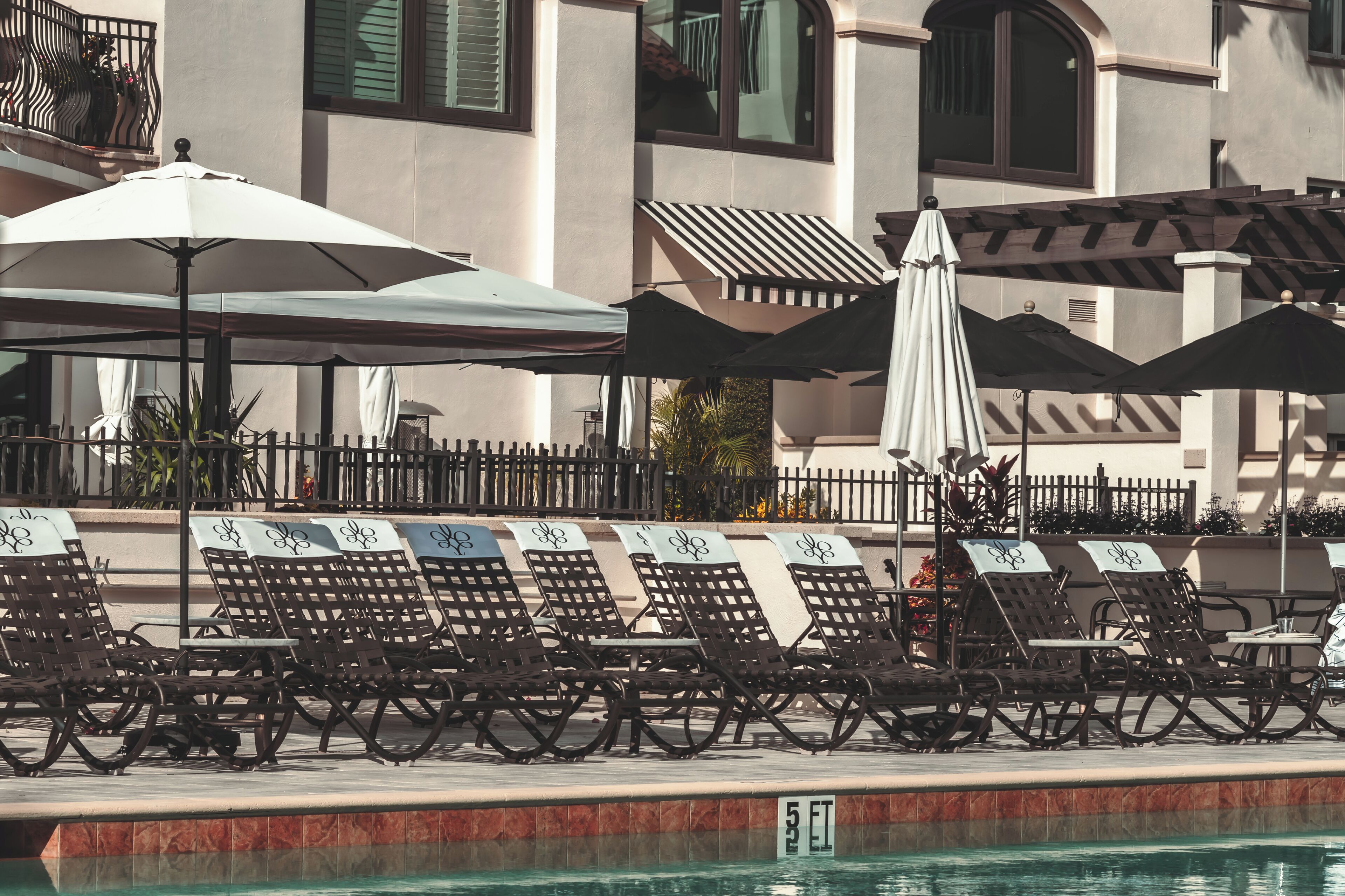 A poolside area with lounge chairs and umbrellas, a tiled pool, and a building with balconies and windows in the background.
