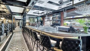 A modern bar area with a long counter, black upholstered bar stools, and a view of the outdoors through large windows.