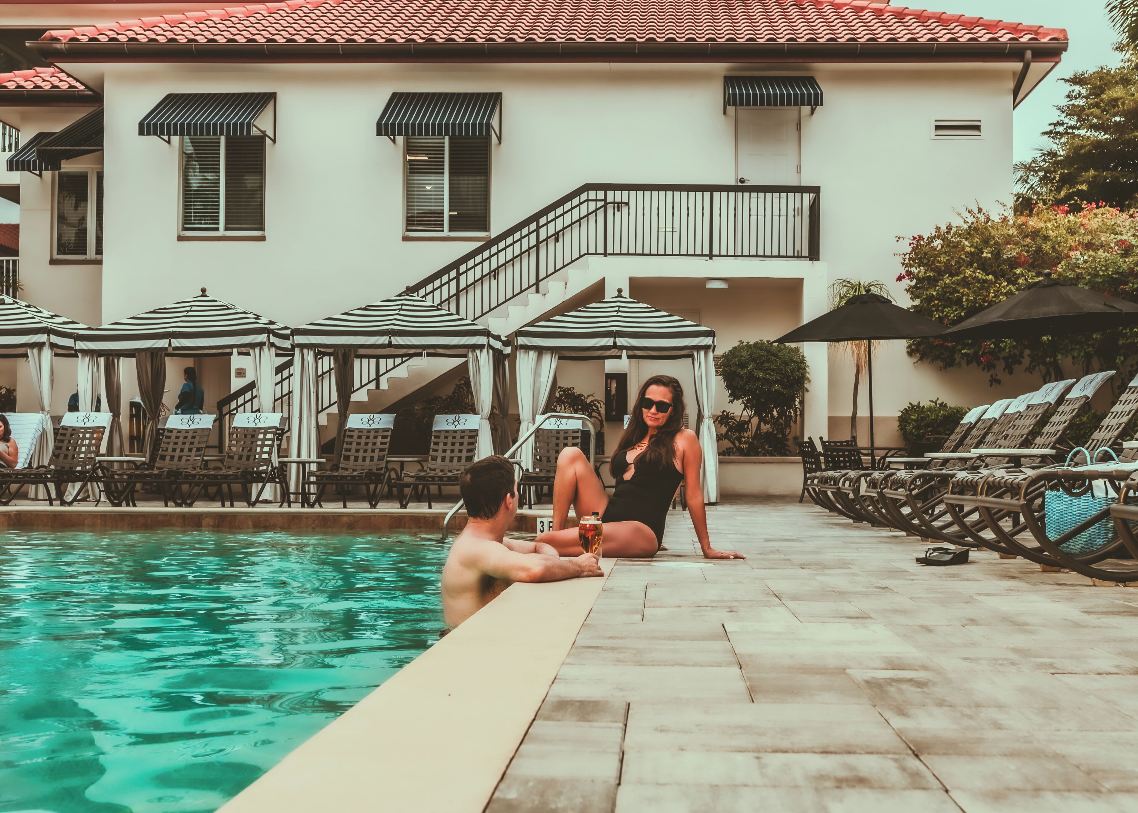 Zona de piscina con sillones de descanso y sombrillas, un edificio con techo de tejas y ventanas, y una escalera que conduce a un nivel superior.