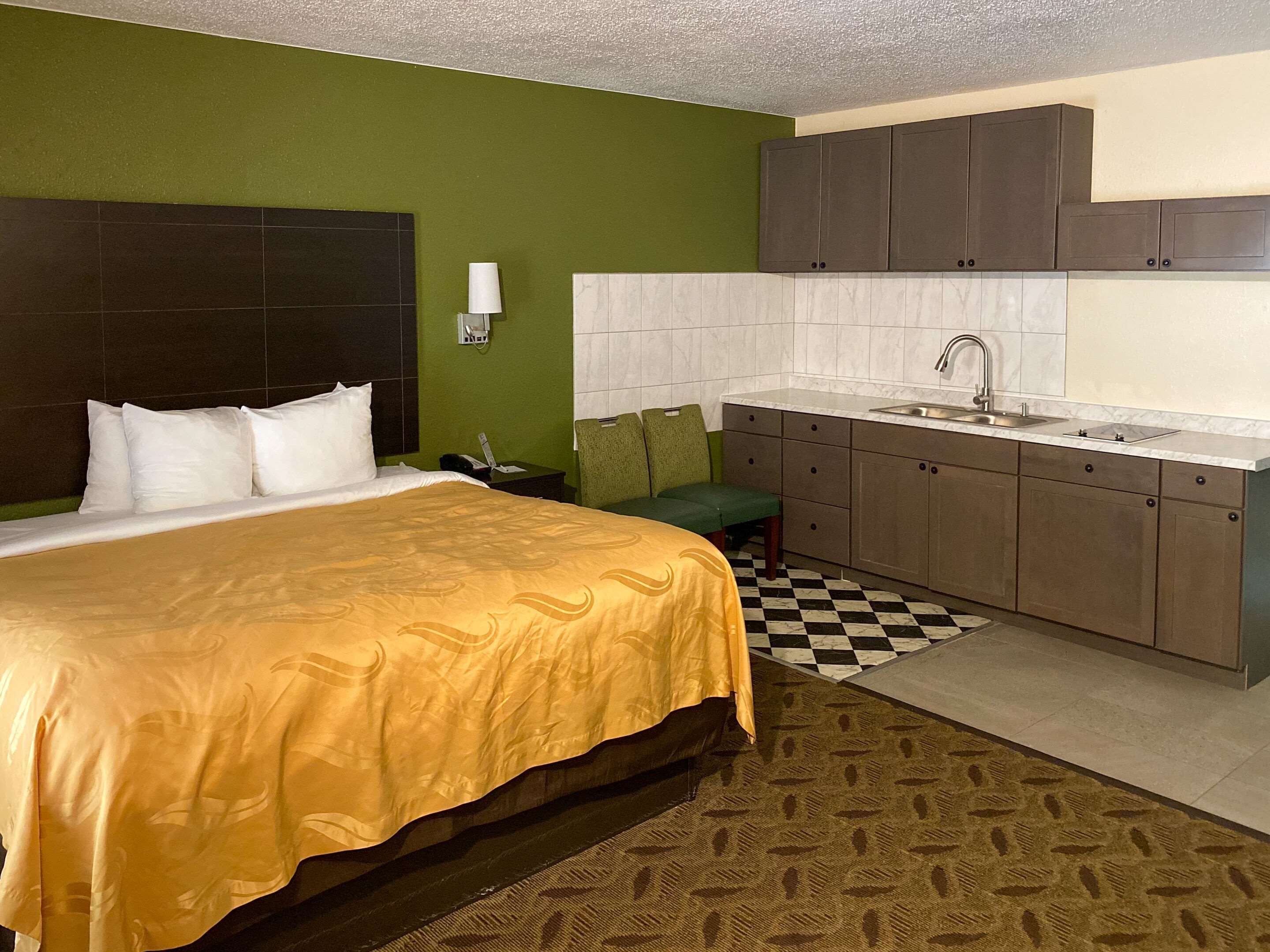 A hotel room with a bed, a green wall, a checkered floor, a kitchen area with a sink and cabinets, and a telephone on the nightstand.