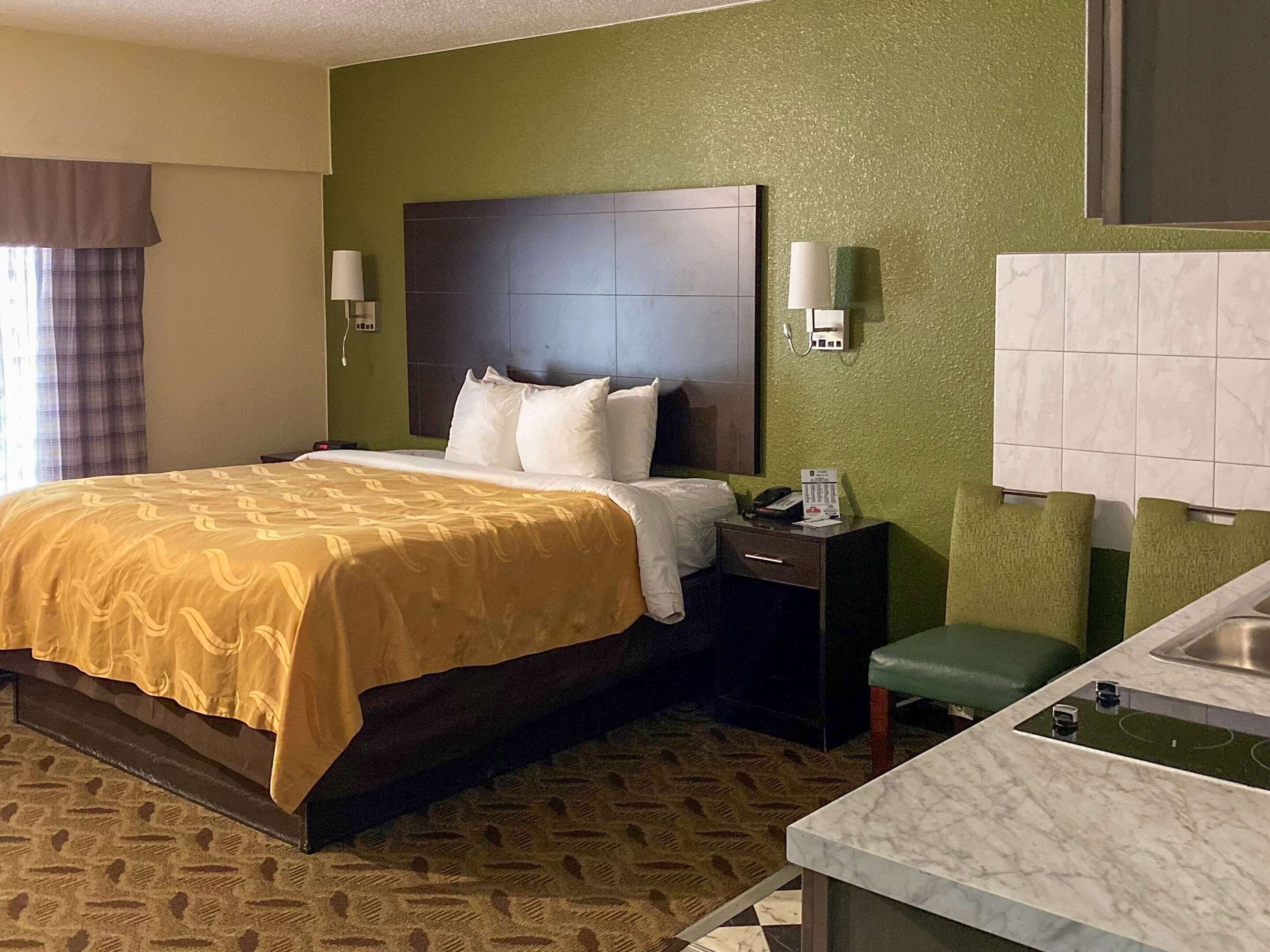 A hotel room with a bed, bedside table, lamp, and a small green stool.