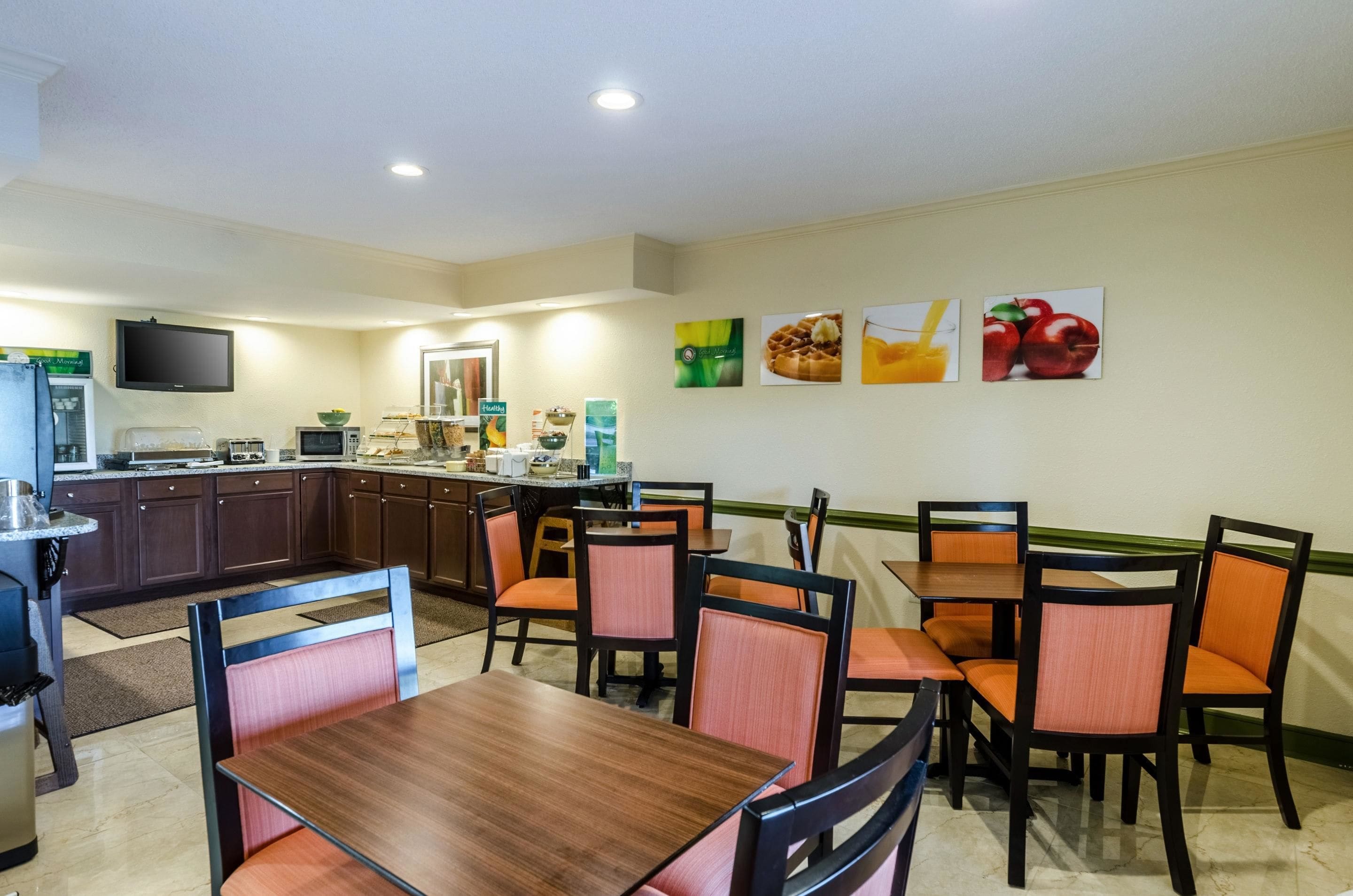 Un comedor con mesas de madera y sillas con cojines naranja, una cocina con refrigerador y televisor de pantalla plana, y decoración mural con representaciones de alimentos.