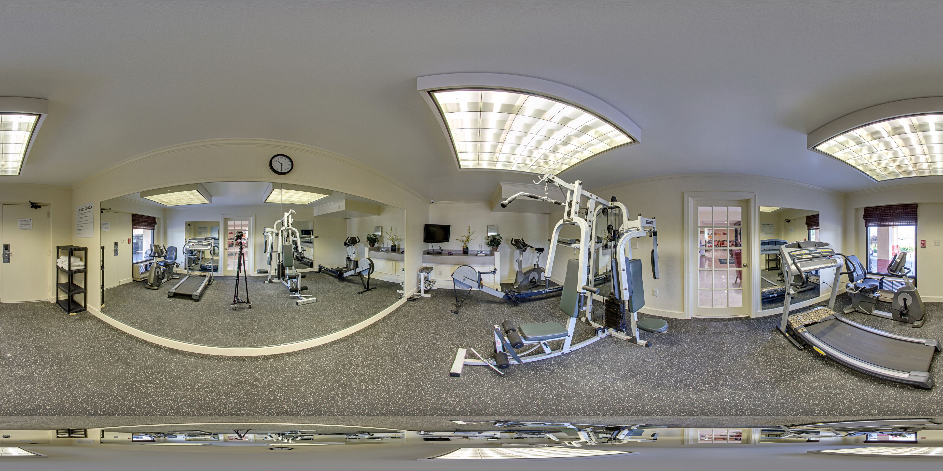 Una vista panorámica de un gimnasio con diversos equipos de ejercicio, un reloj en la pared y un techo con tragaluces.