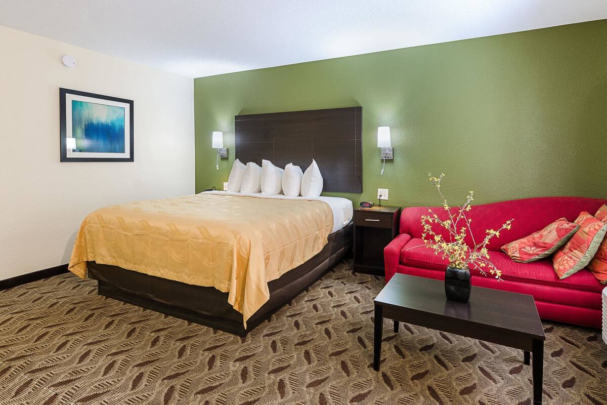 A hotel room with a bed, a red sofa, a small table with a vase, and a framed picture on the wall.