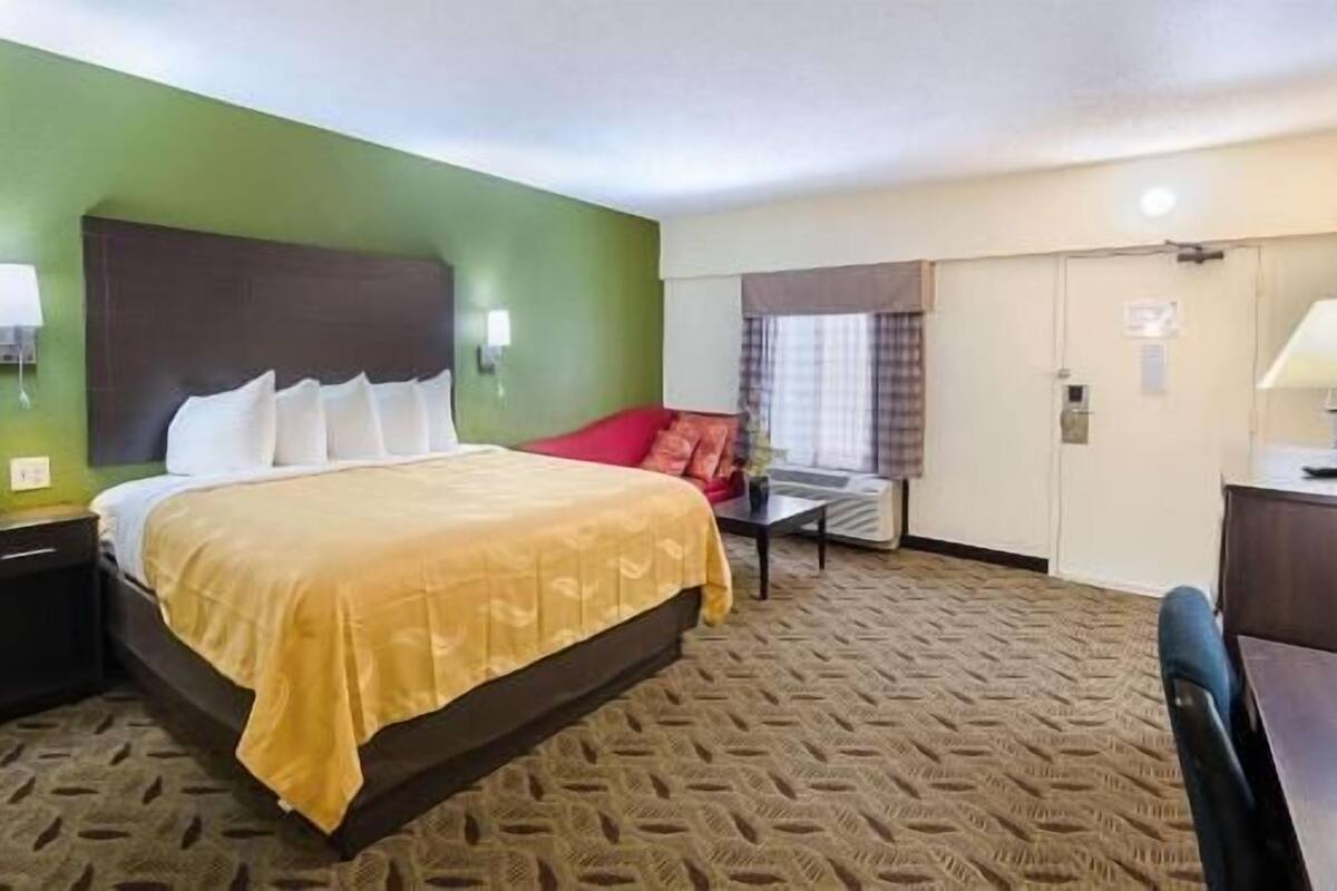 A hotel room with a bed, a desk, a chair, a window with curtains, and a door.