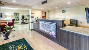 A reception area with a reception desk, a waiting area with a red chair, and a reception counter with a logo. - Quality Inn Tanglewood (Roanoke)