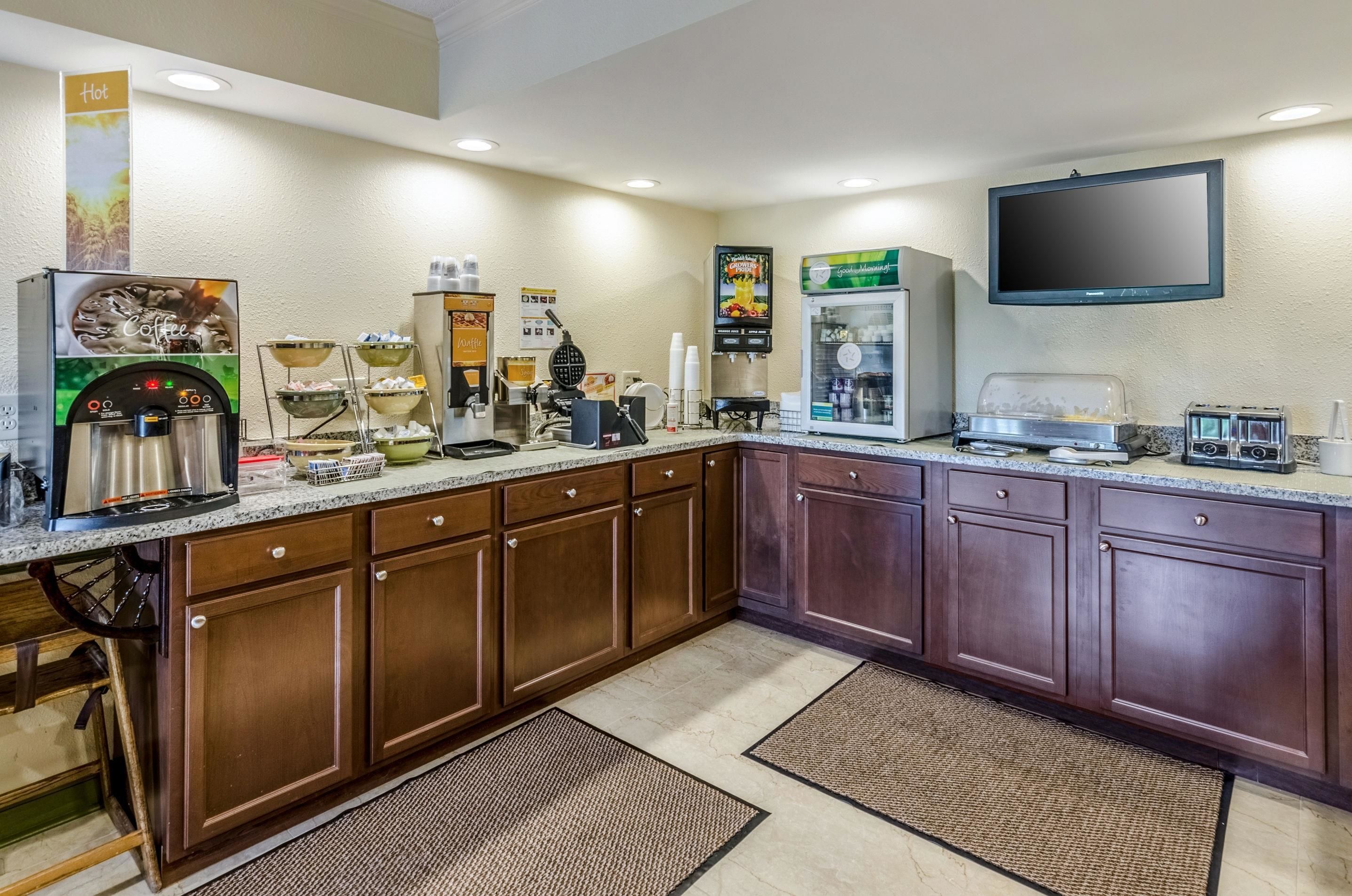 Una cocina de hotel con una máquina de café, un mostrador de alimentos, un dispensador de bebidas y un televisor montado en la pared.
