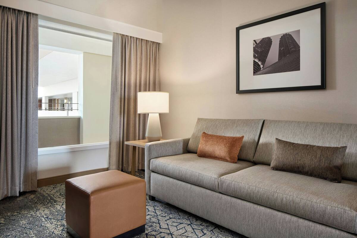 Ein Hotelzimmer mit grauem Sofa, einem kleinen Hocker, einem Beistelltisch mit Lampe und einem gerahmten Bild an der Wand.