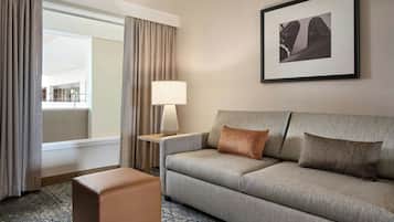 A hotel room with a grey sofa, a small ottoman, a side table with a lamp, and a framed picture on the wall.