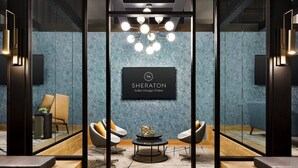 A modern hotel lobby with a glass partition, a sign for Sheraton Suites Chicago O'Hare, two armchairs, a round table, and a decorative globe. - Sheraton Suites Chicago O'Hare (Rosemont)
