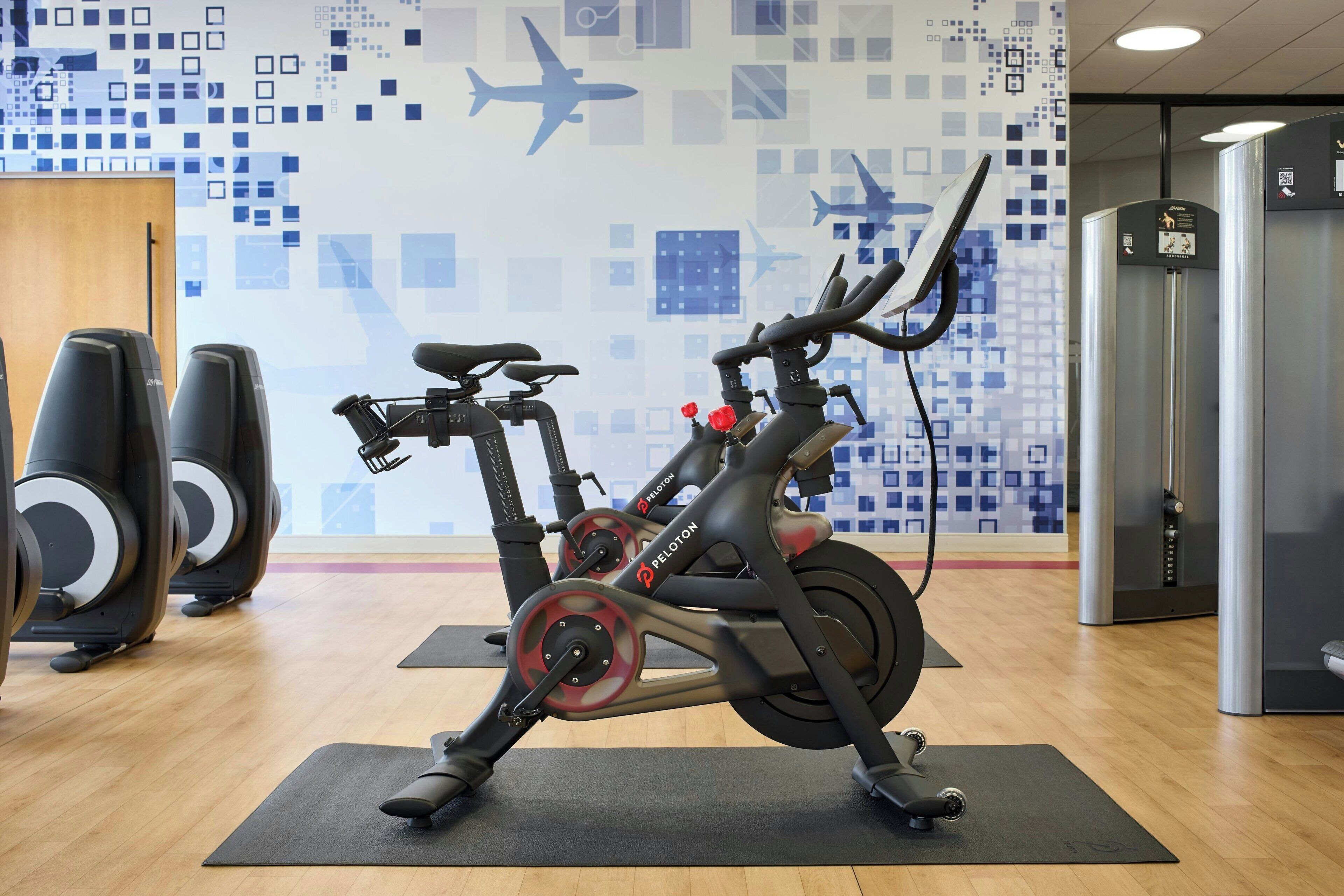 A Peloton exercise bike in a gym with a cityscape wall mural, wooden flooring, and a door to a changing room.