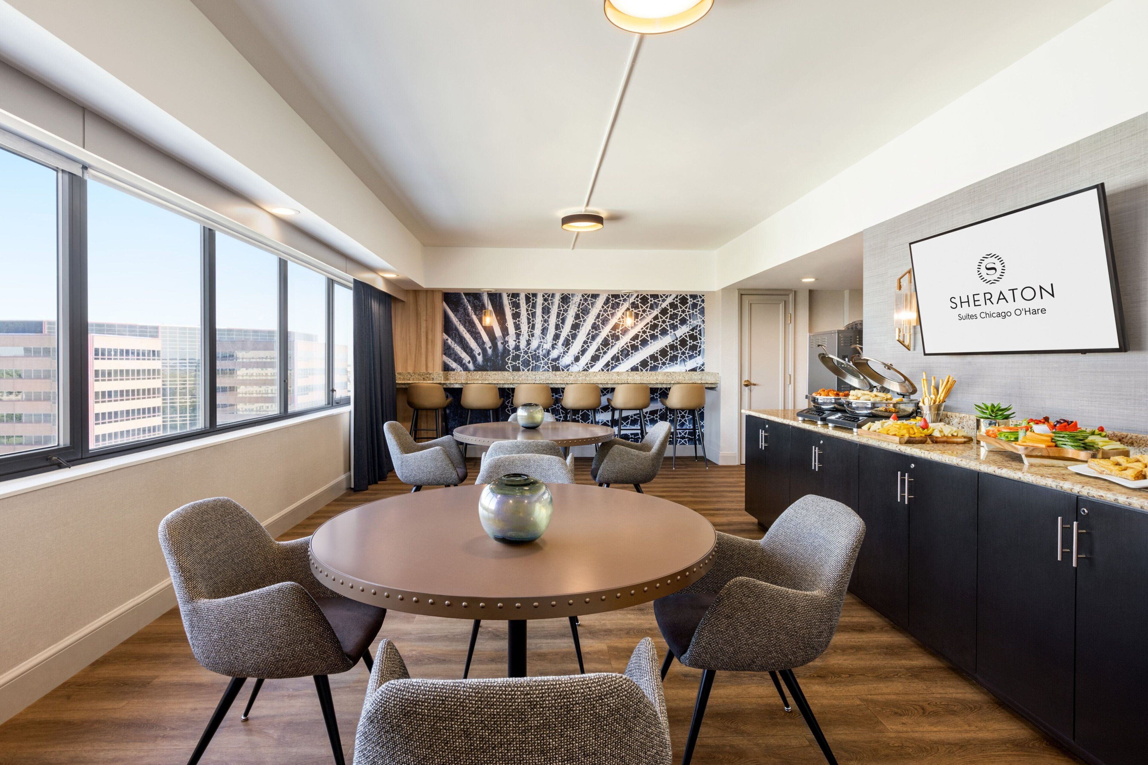 Habitación de hotel moderna con comedor, un ventanal con vistas a la ciudad y el logotipo de Sheraton en la pared.