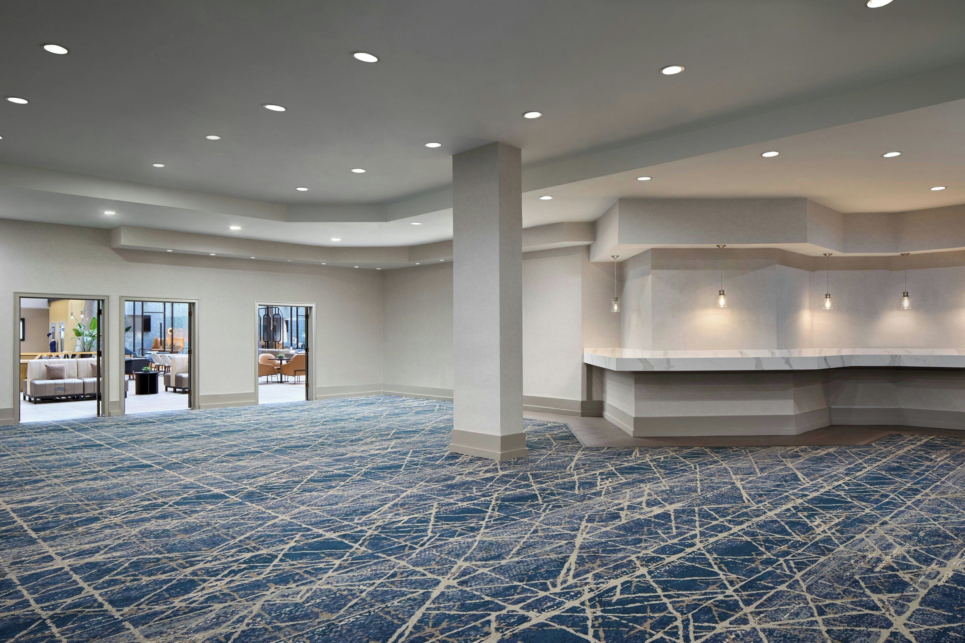 A spacious hotel lobby with a blue patterned carpet, recessed lighting, and a seating area visible through glass doors.