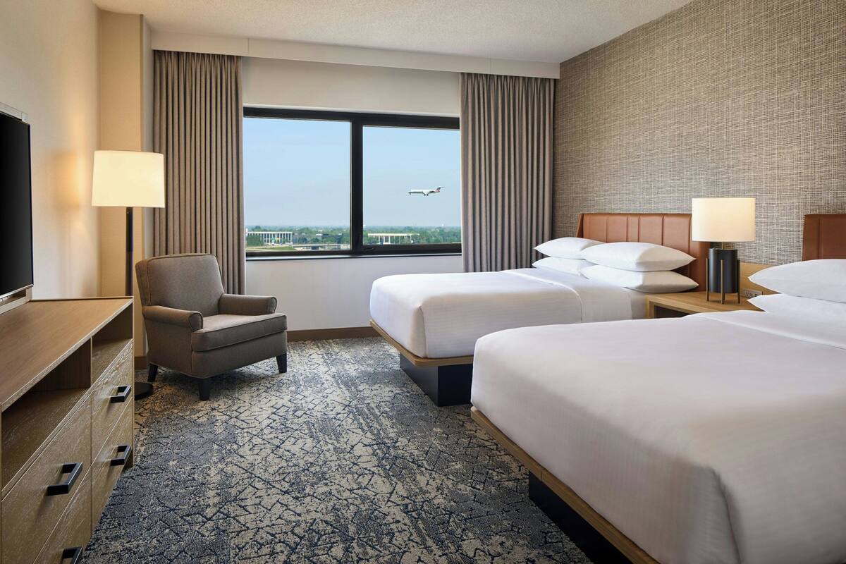 Ein Hotelzimmer mit großem Fenster, zwei Betten, einem Sessel, einem Schreibtisch und einem Fernseher.