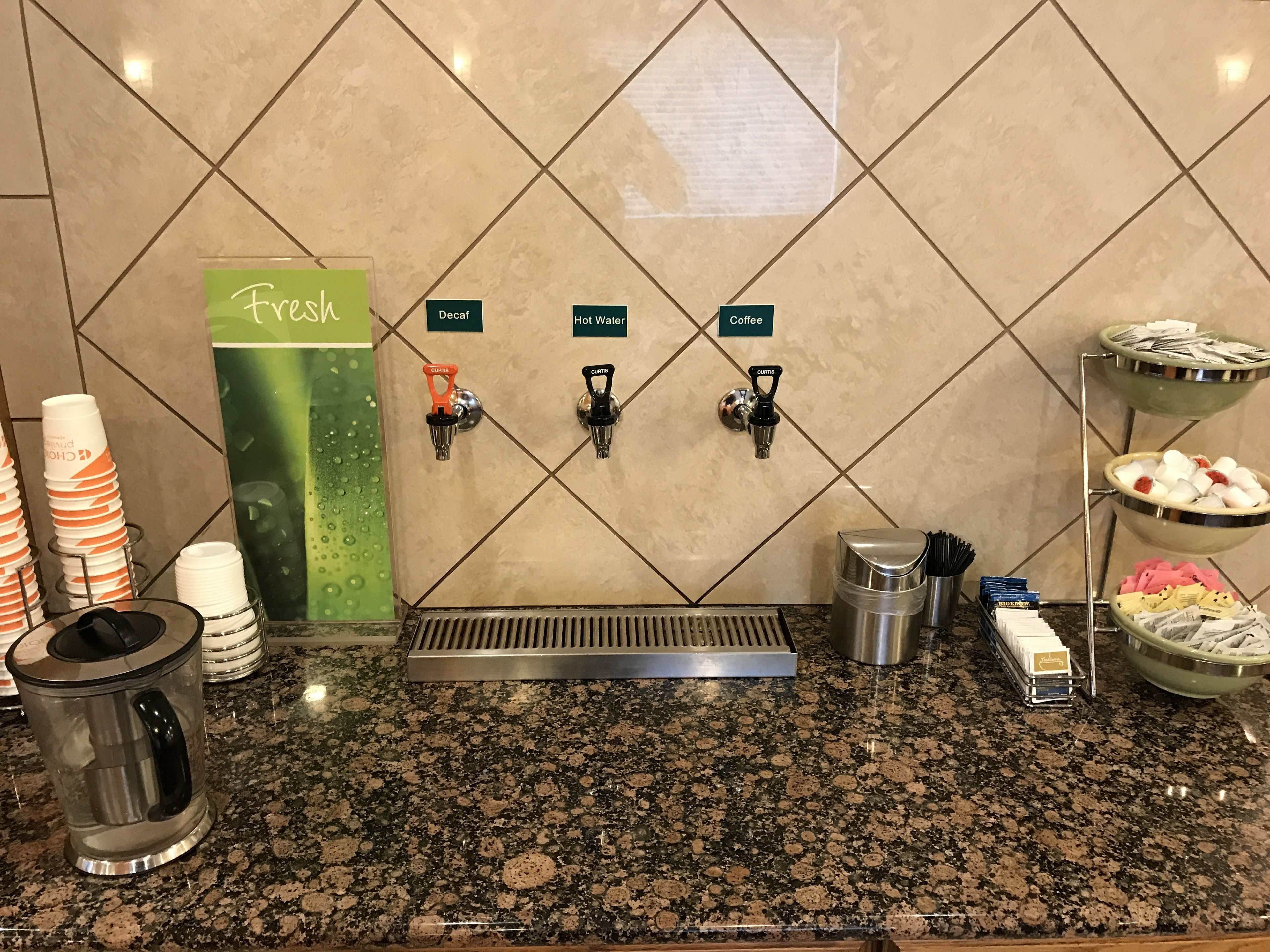 A coffee station with dispensers for decaf, hot water, and coffee, a fresh sign, and a bowl of assorted snacks.