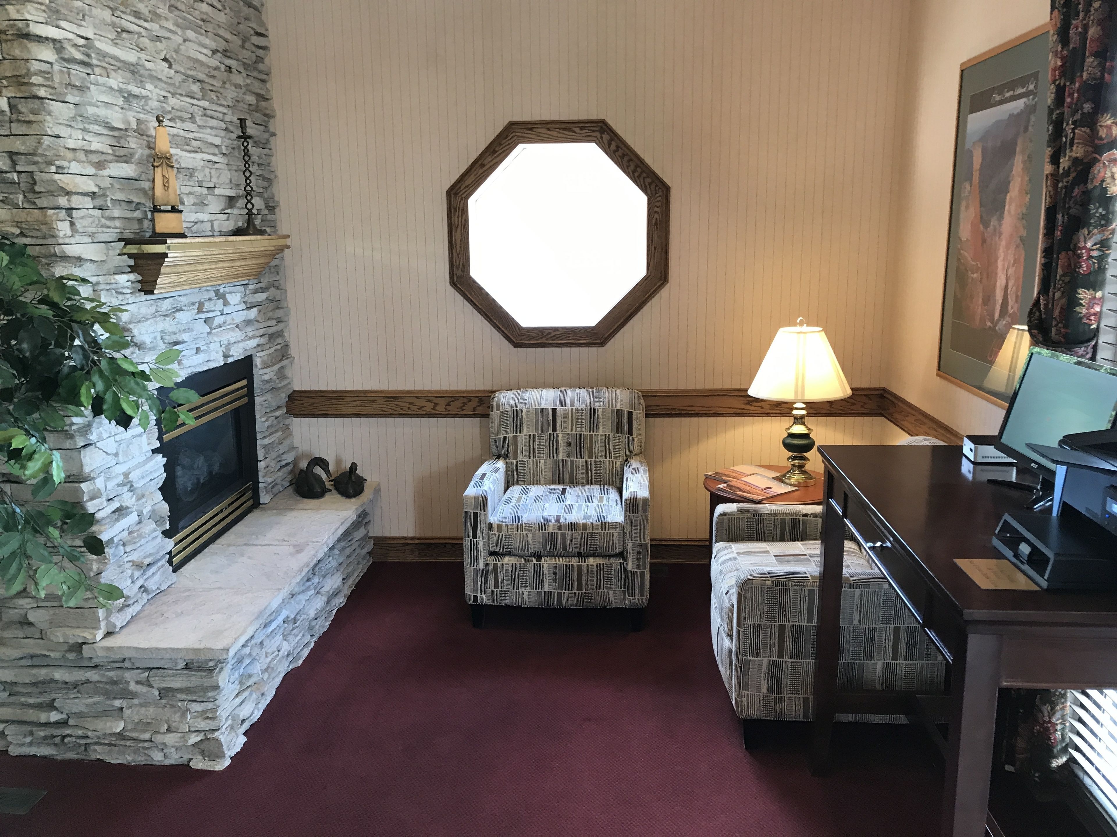 A room with a stone fireplace, two patterned armchairs, a desk with a lamp, and a large window.