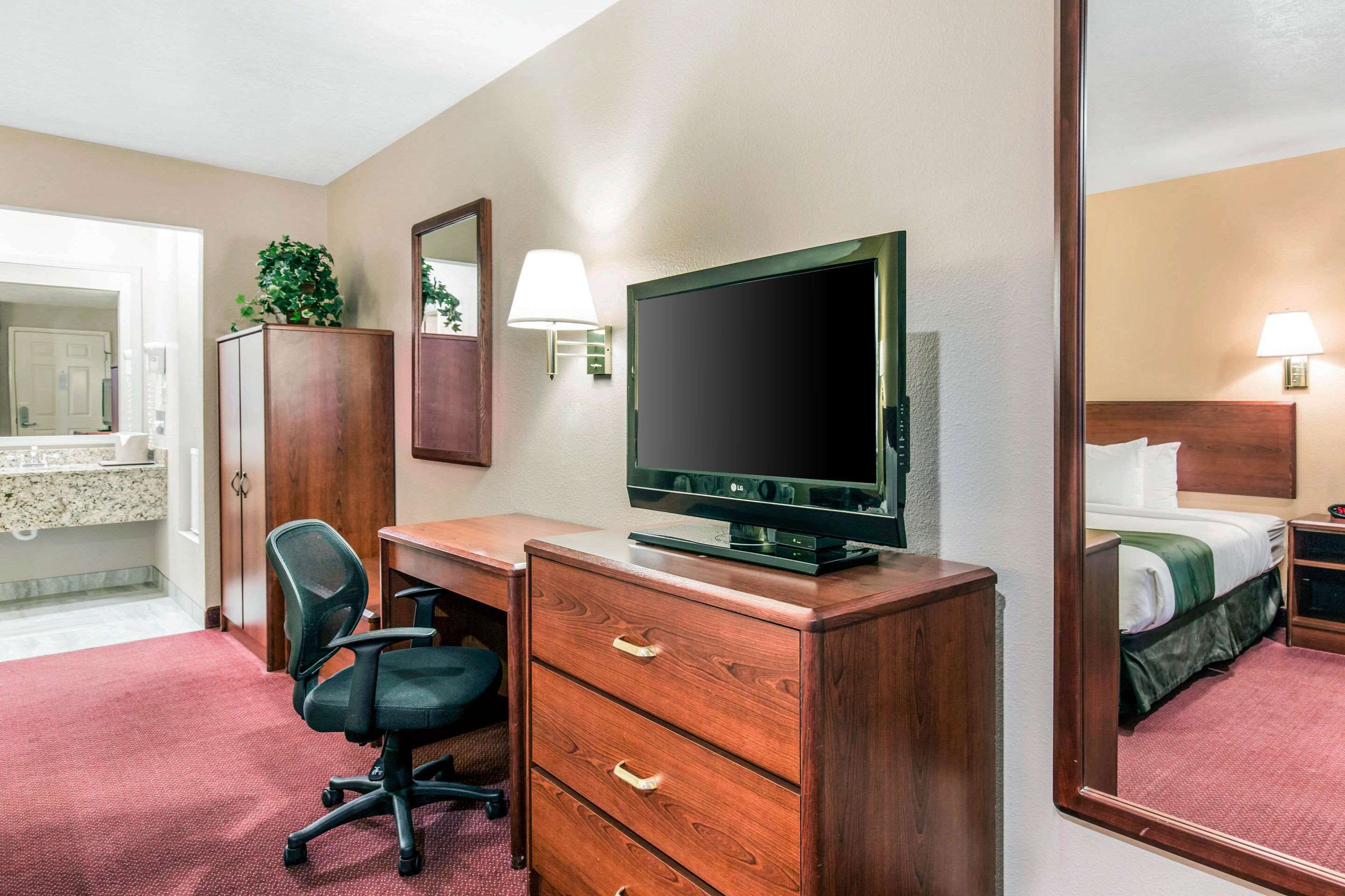 A hotel room with a wooden desk, a television, a mirror, a bed, and a bathroom visible in the background.