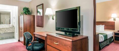 A hotel room with a wooden desk, a television, a mirror, a bed, and a bathroom visible in the background.