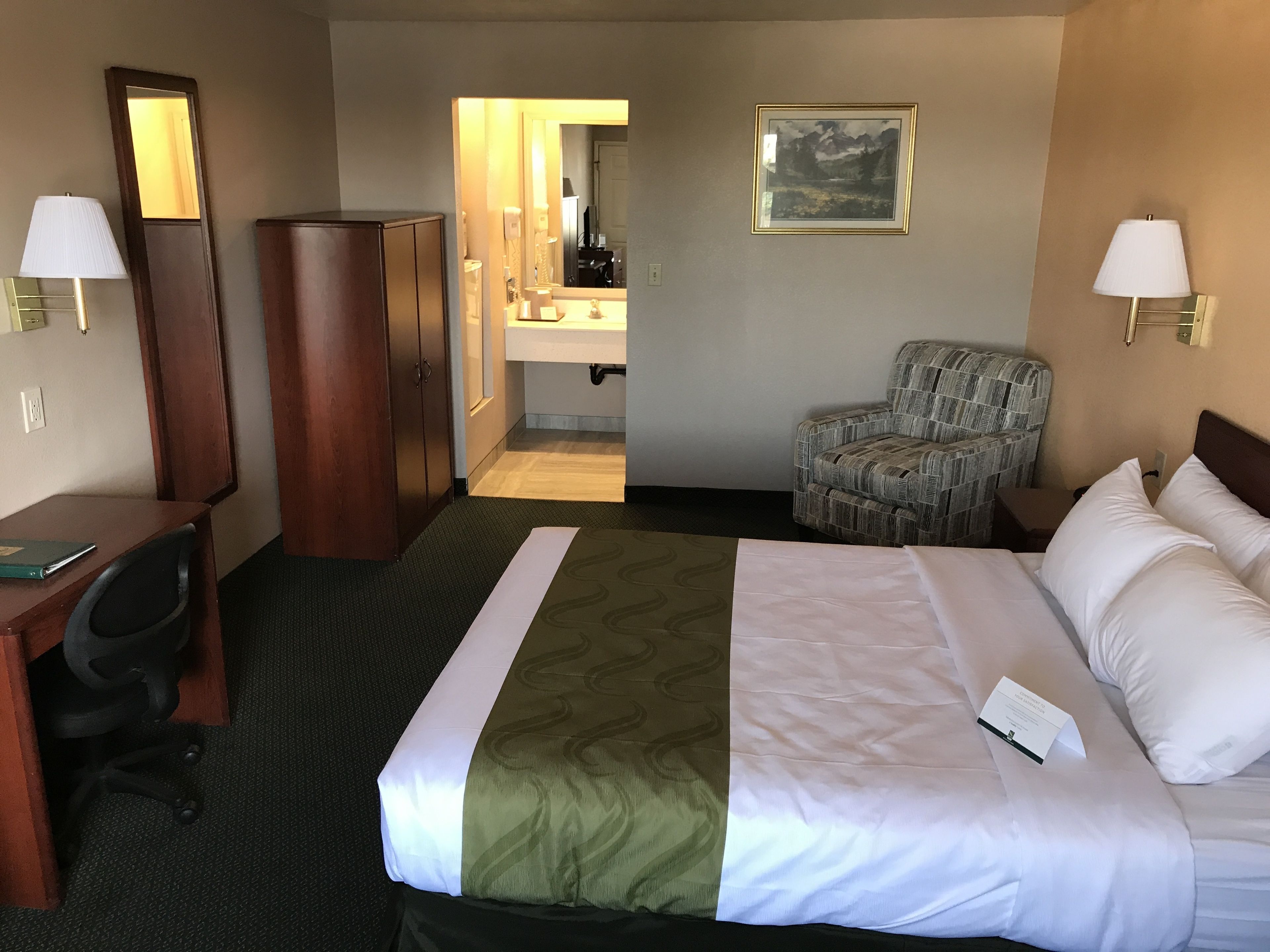 A hotel room with a bed, a desk, a chair, a wardrobe, a bathroom visible through an open door, and a framed picture on the wall.